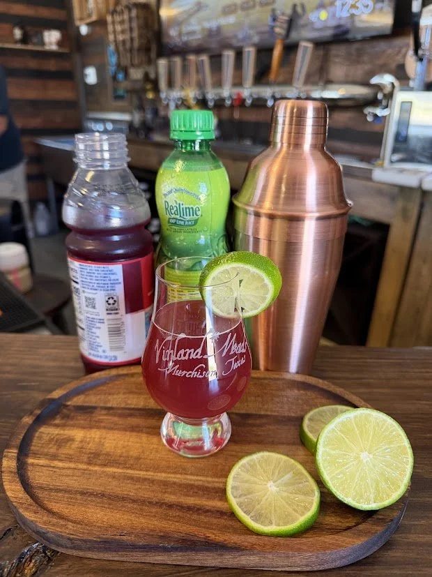 A cocktail glass with a pink drink and a lime wedge garnish on a wooden tray. Behind the tray are bottles including a red beverage, a green bottle of Realime, and a shiny copper cocktail shaker in a bar setting.