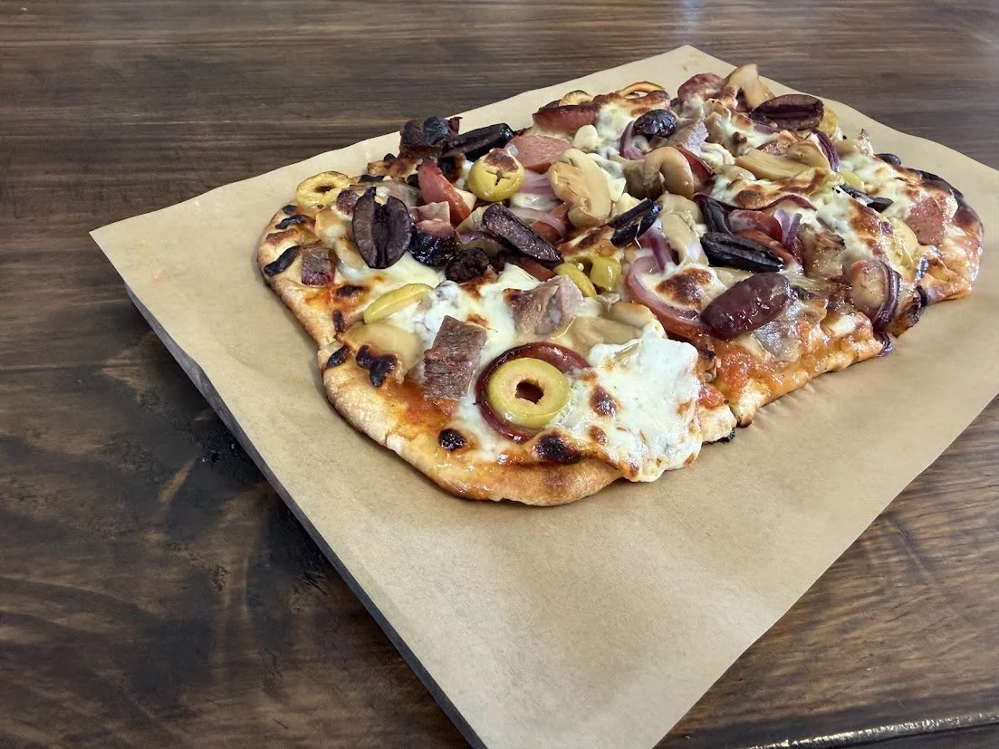 A rectangular flatbread style pizza with a variety of toppings including olives, onions, cheese, and meat on a piece of parchment paper on a wooden table.