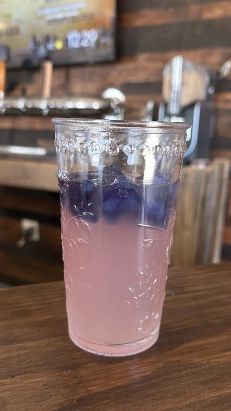 A tall glass with a pink drink topped with blueberries on a wooden table.