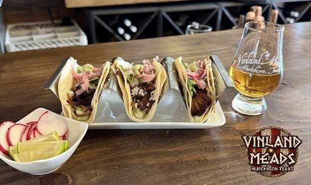 Three tacos with various fillings on a white platter, a small bowl of sliced radishes and lime wedges, and a glass of mead on a wooden bar counter.