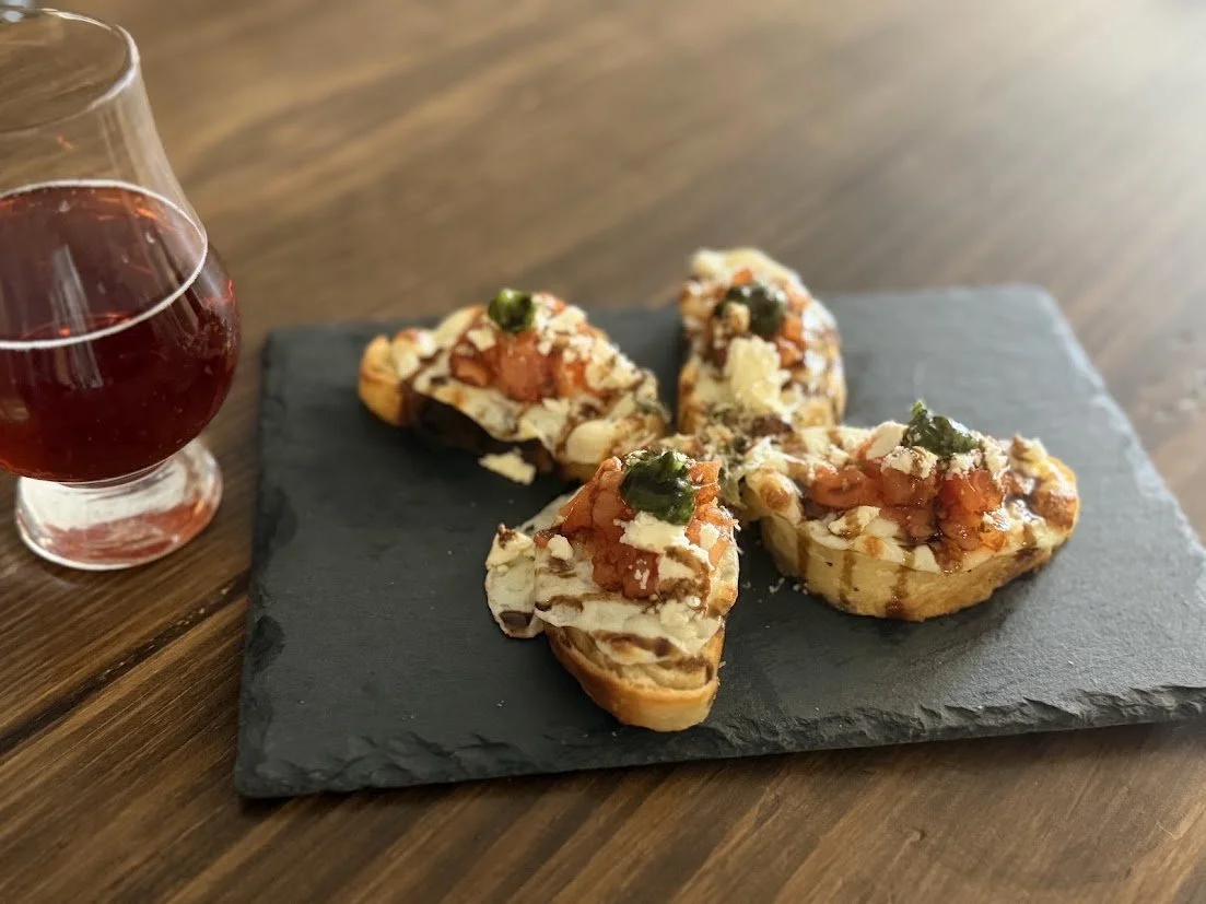 Four slices of bruschetta with tomato, cheese, and herbs on a black slate platter, with a glass of red mead on a wooden table.