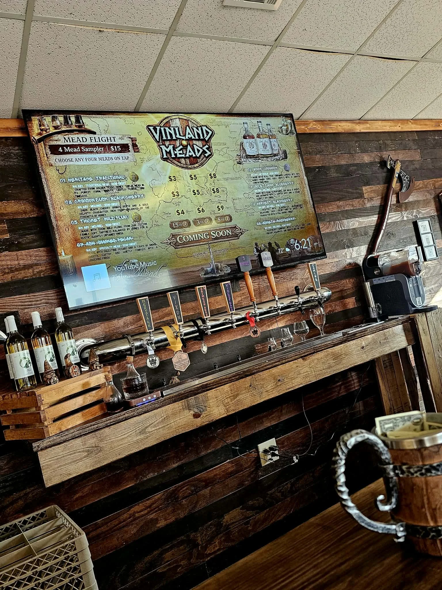 A digital menu board displaying various meat options and prices at a bar or restaurant, with beer taps and bottles of alcohol below it and a wooden wall background.