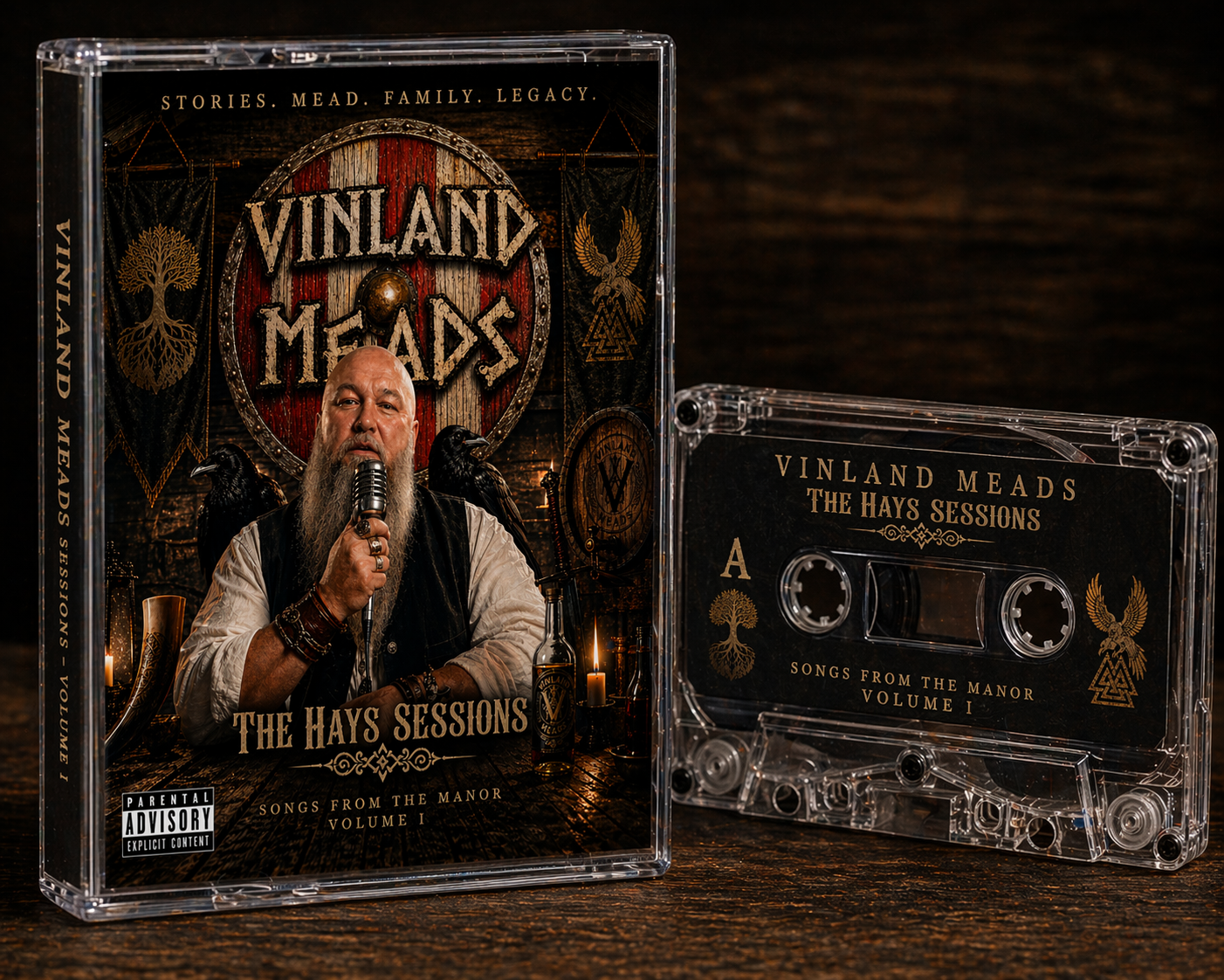 A music album cover titled "Vinland Meads" with the subtitle "The Hays Sessions" and the description "Songs from the Manor Volume 1". It features a man with a long beard holding a microphone, wearing rings and bracelets, sitting at a table with candles and alcohol bottles. The background has medieval and Norse symbols, banners, and a large shield with the album title.