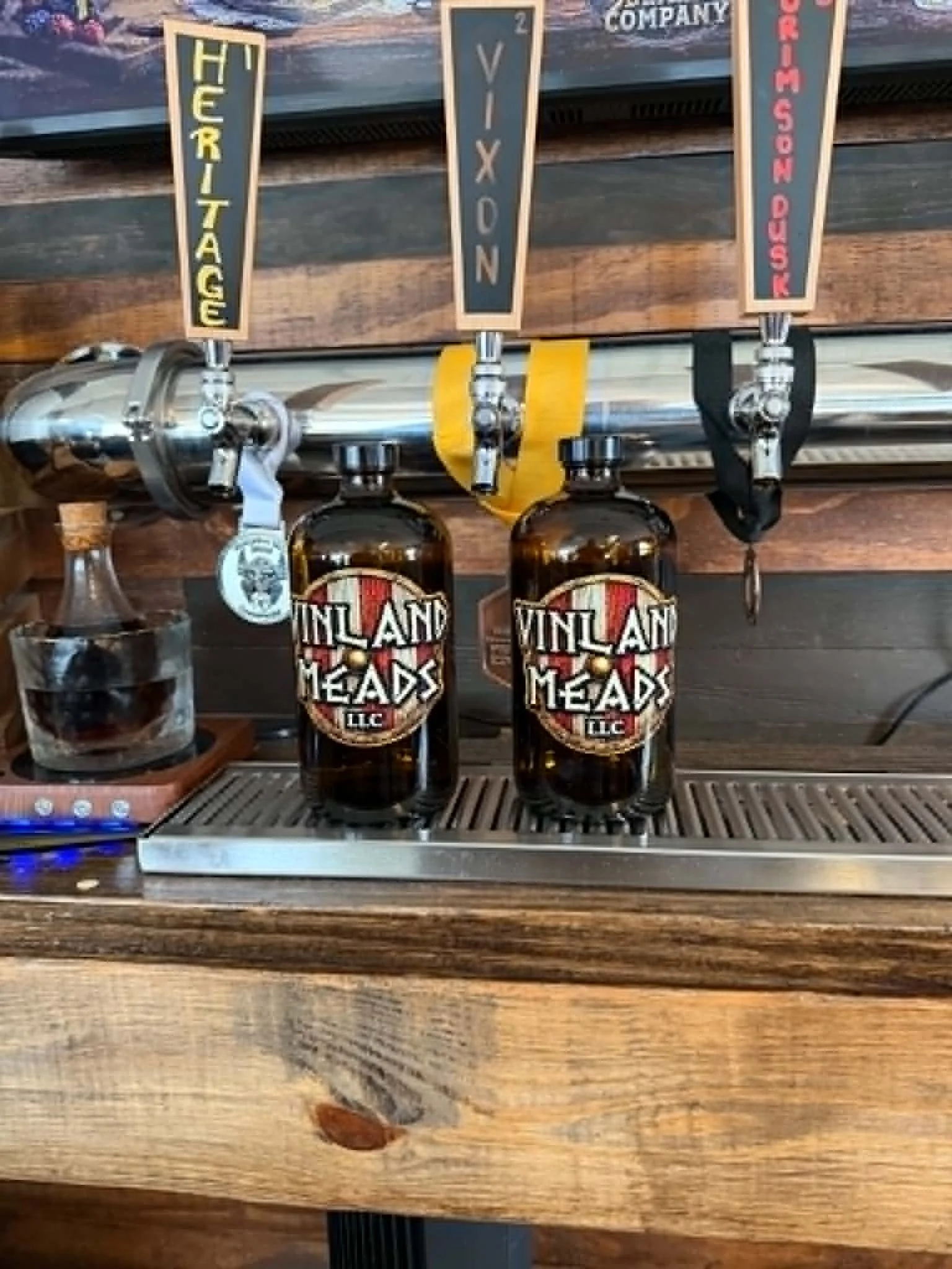 Three mead taps with black and red labels reading 'VINLAND MEADS,' mounted on a shiny metal mead tower, with a mead growlers in the foreground, on a wooden bar counter.