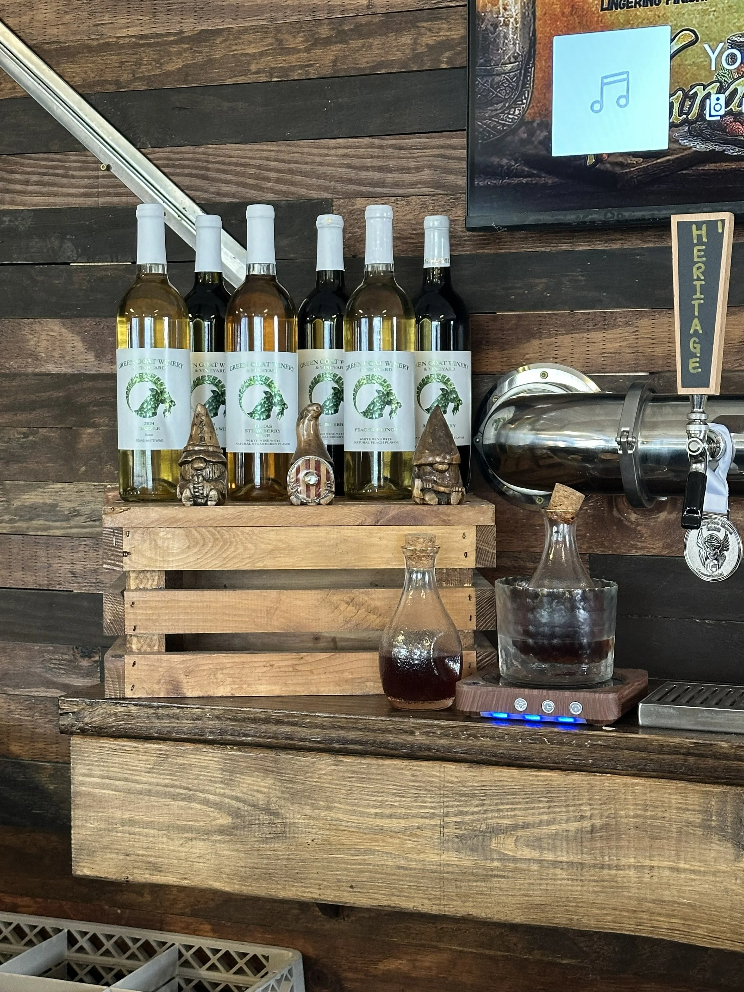 Display of six bottles of white and red wine on a wooden shelf with decorative gnome figurines, a glass container with dark liquid, and a small sign that says 'HERITAGE.' A digital scale and a glass of dark liquid are on the counter below.