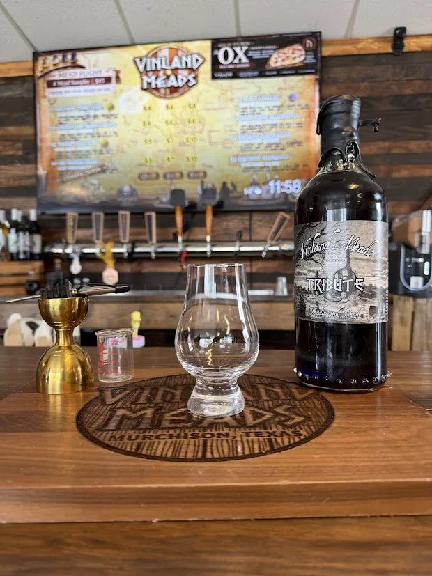 Bar counter with a empty mead glass, a gold shot measuring glass, a dark mead bottle with a label that says "Tribute", and a small glass. In the background, a menu board lists food items, and tap handles are mounted on a wooden wall.