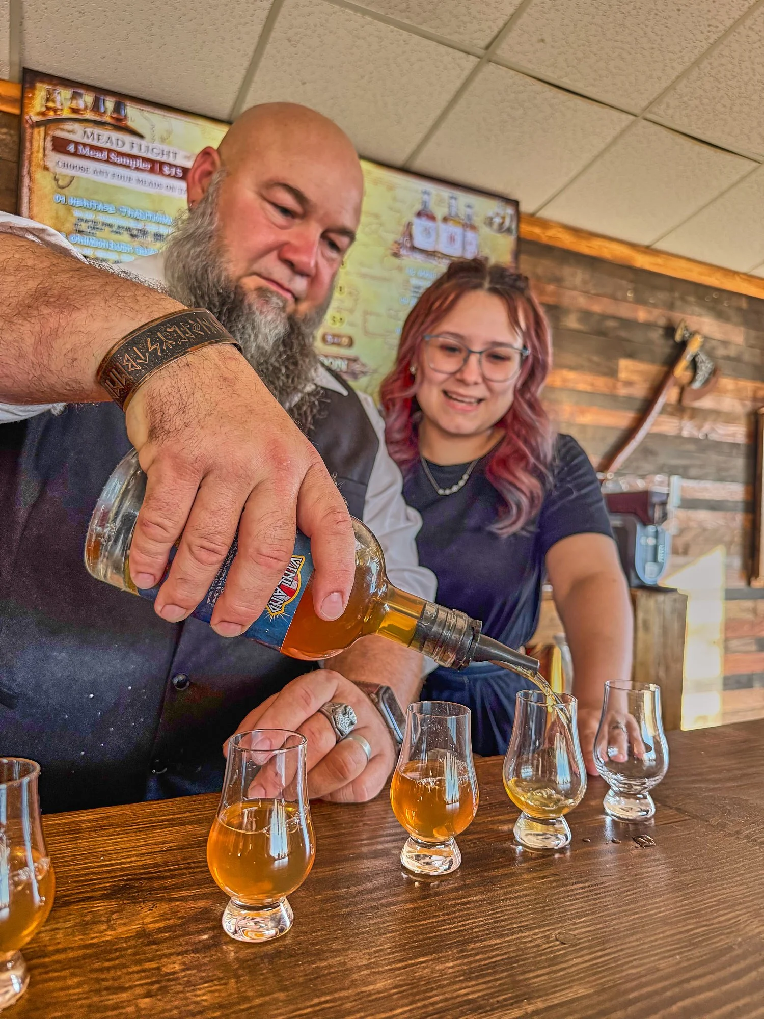A man with a beard and a woman with glasses pour a drink from a bottle into a glass at a wooden table with several glasses filled with a golden beverage in a rustic restaurant setting.