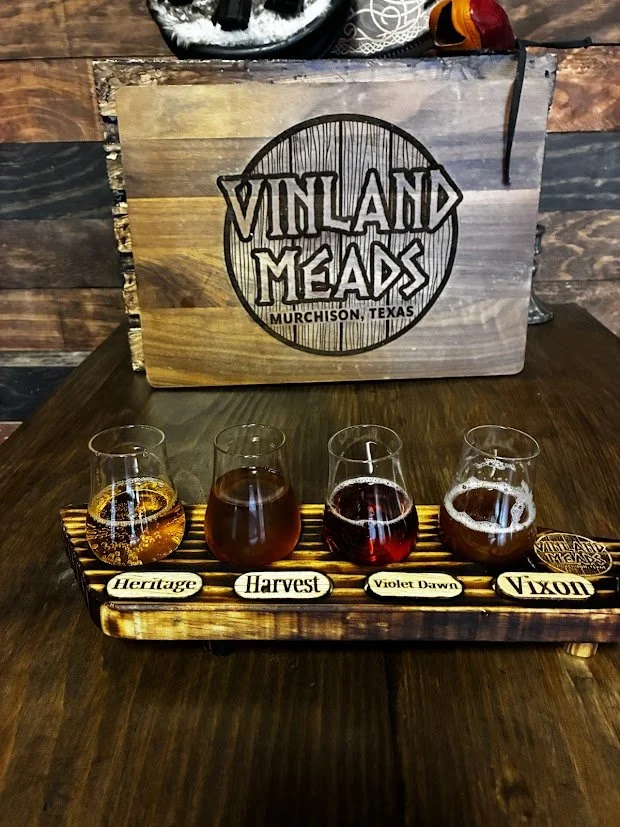 Four glass mead tasters on a wooden flight with labels: Heritage, Harvest, Violet Dawn, Vixon; behind a wooden sign reading 'Vinland Meads Murchison, Texas'.