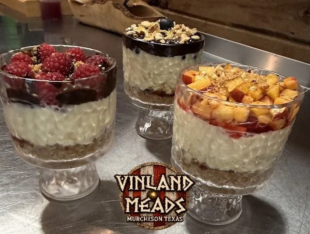 Three glass dessert cups filled with layered desserts topped with berries, chocolate, and fruit, on a metal surface with a Vinland Meads logo.