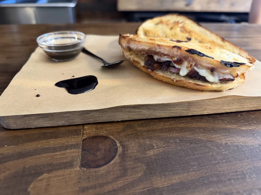 A grilled cheese sandwich with brisket  inside on a wooden serving board, accompanied by a small jar of balsamic glaze and a small bowl of dipping sauce, set on a brown paper liner.
