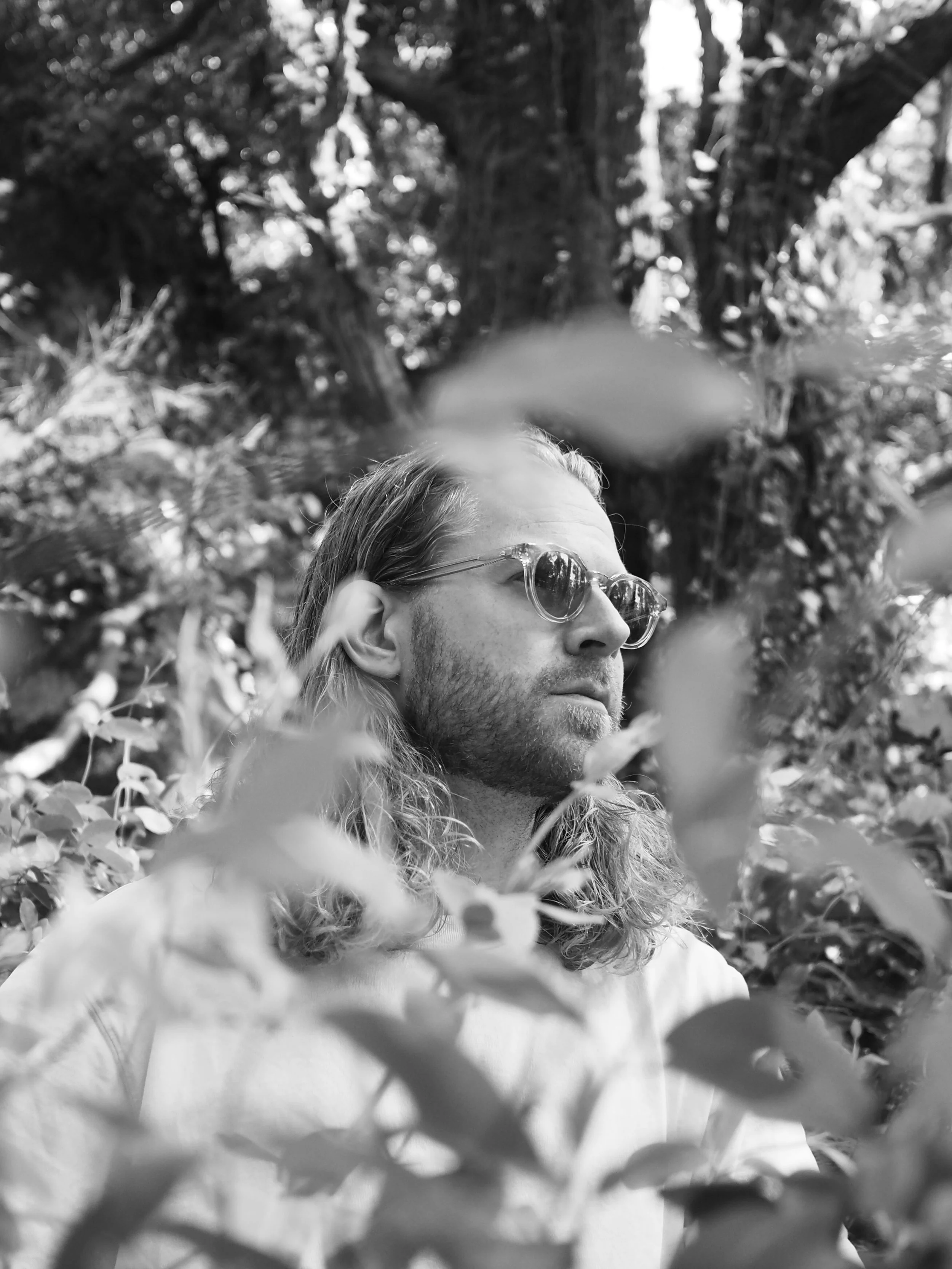 A man with long hair, sunglasses, and a beard, looking to the side in a forest setting with blurred leaves in the foreground.
