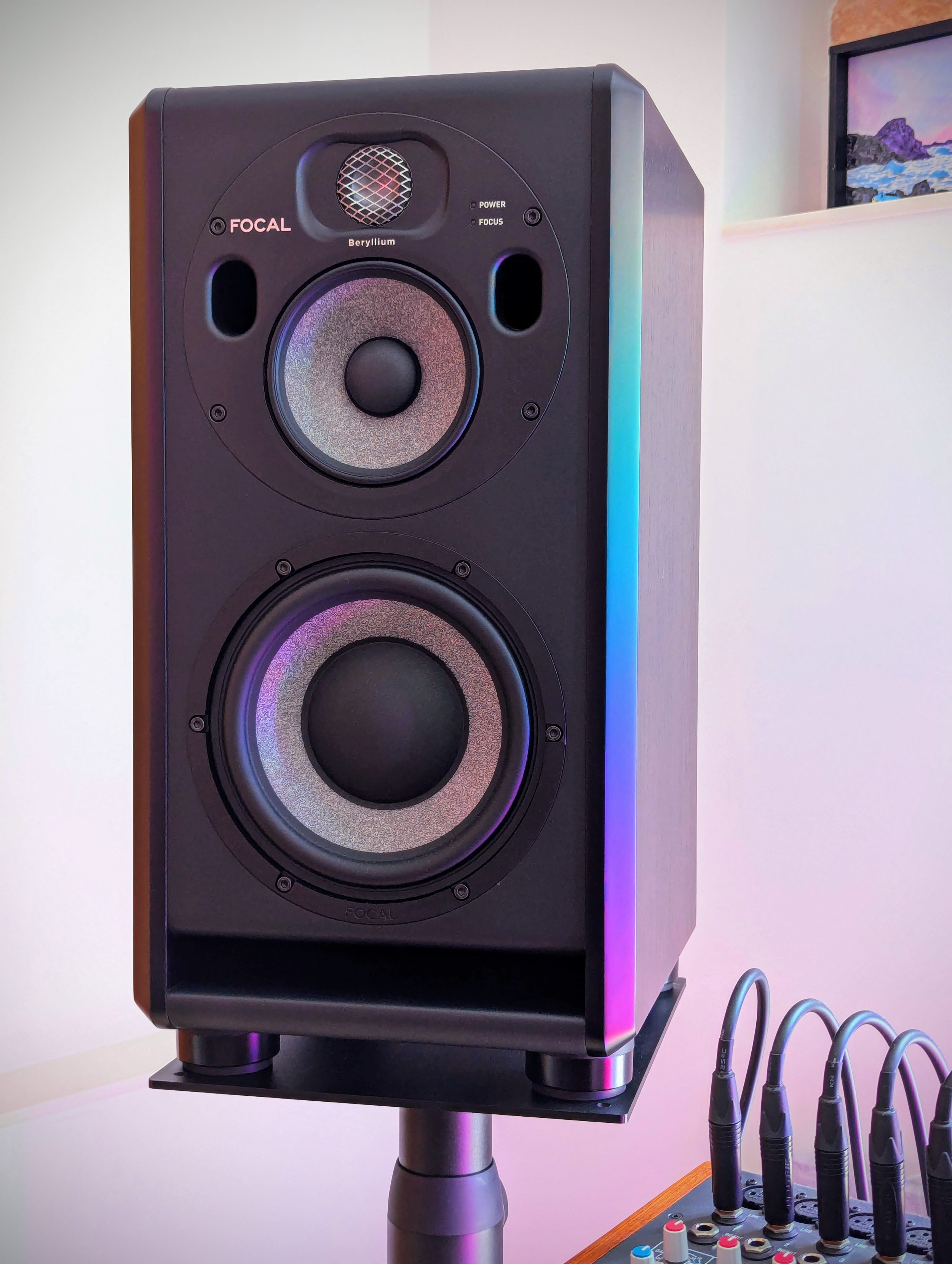 A black Focal speaker with multiple drivers, standing on a stand, with colorful LED lighting on its side, placed next to an audio mixing console.