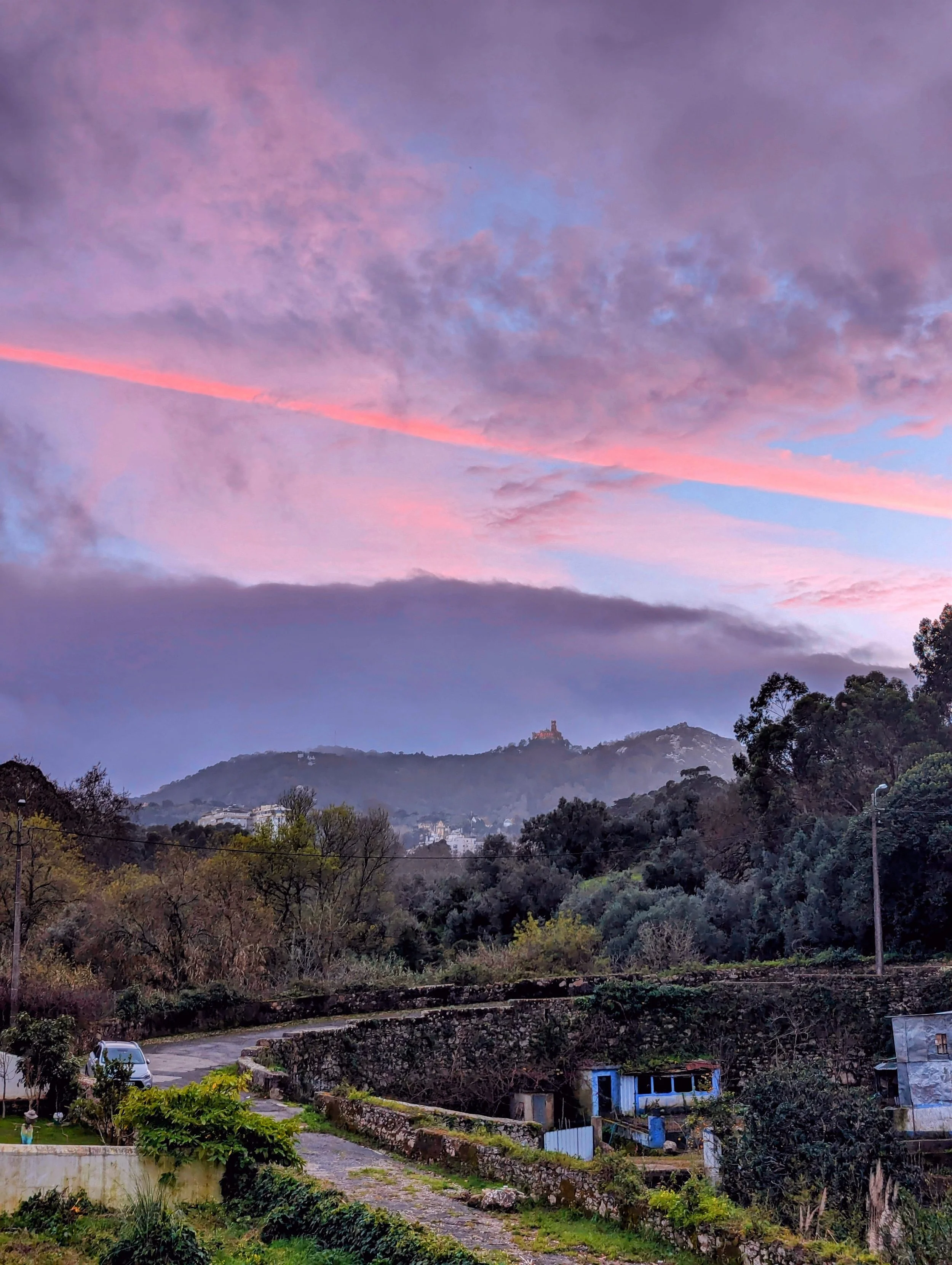 Scenic view of a rural area with small houses, greenery, and a winding road, mountains in the background, and a colorful sunset with pink, purple, and blue clouds.