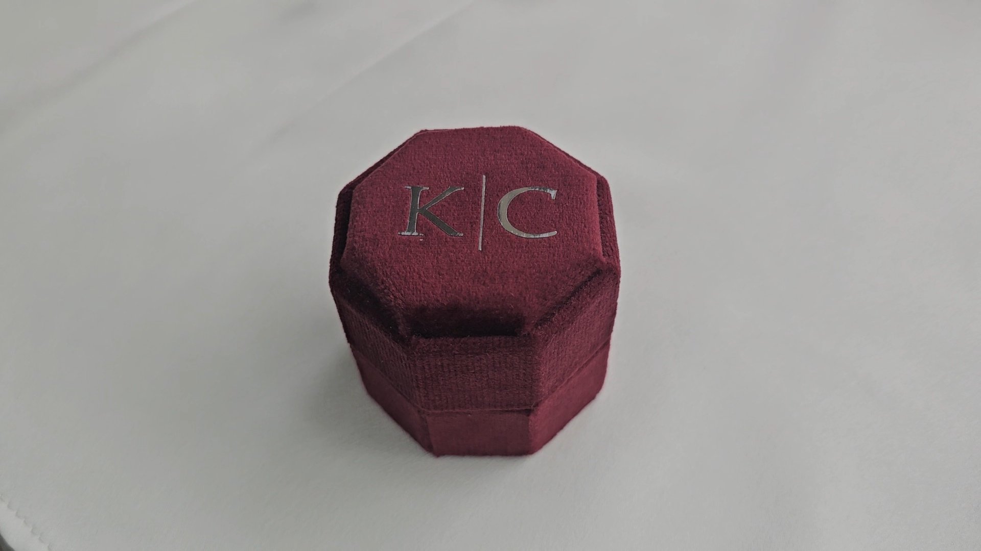 A maroon, hexagonal velvet jewelry box with silver initials "K / C" on top, placed on a white surface.