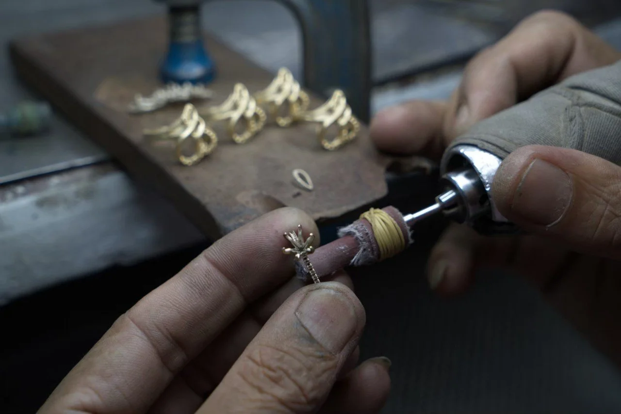 A jeweler uses a rotary tool to polish or engrave a gold jewelry piece, with gold jewelry pieces on a workbench in the background.
