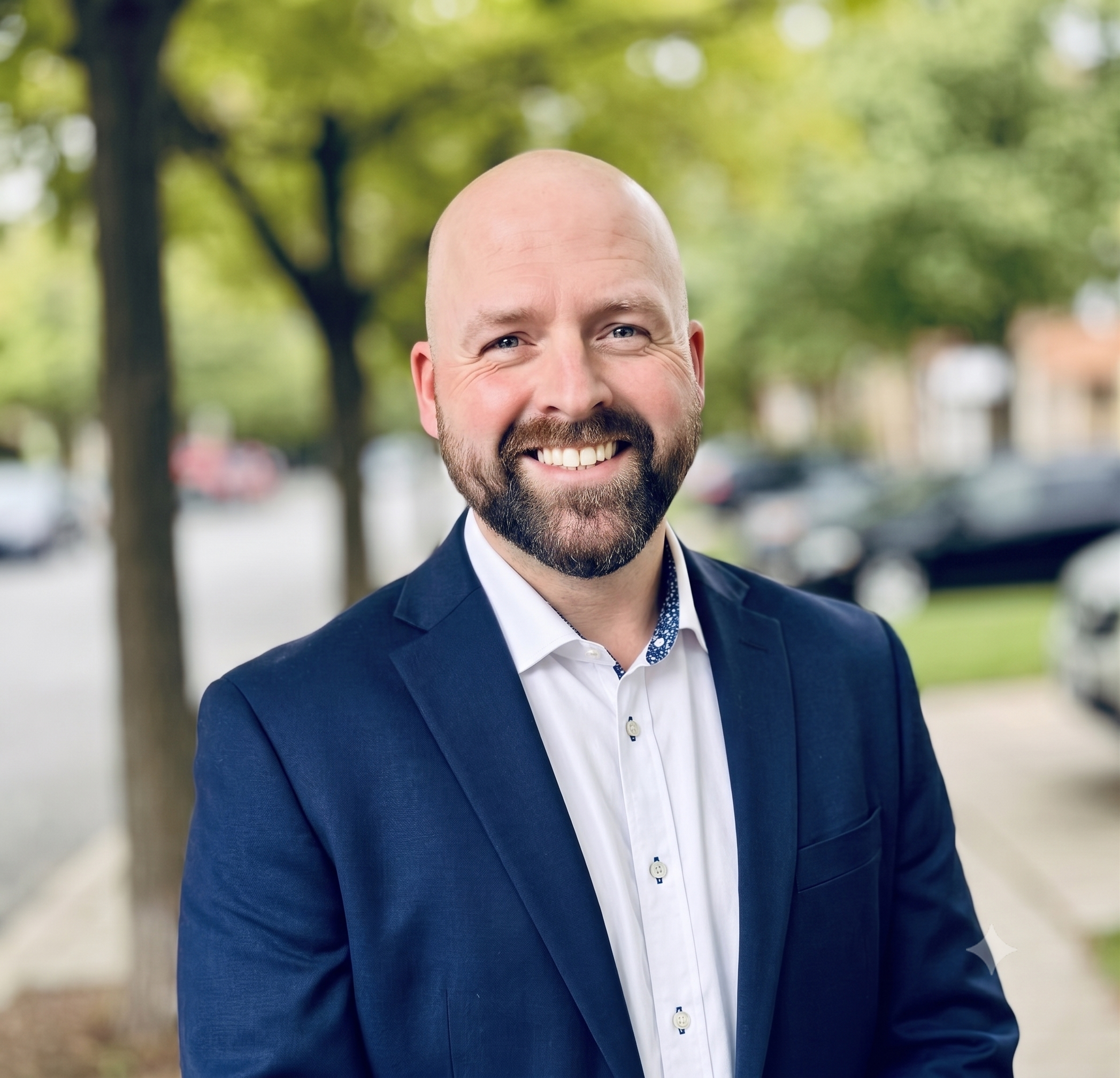 Close-up of a smiling, bald man with a beard, wearing a navy blazer and white shirt, standing outdoors with blurred green trees in the background.