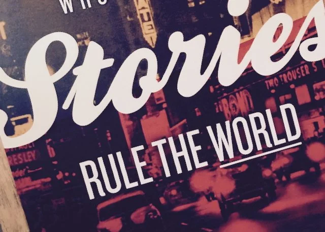 Close-up of a poster with large text saying "Stories Rule the World" over a background of a city scene with neon lights and signs.