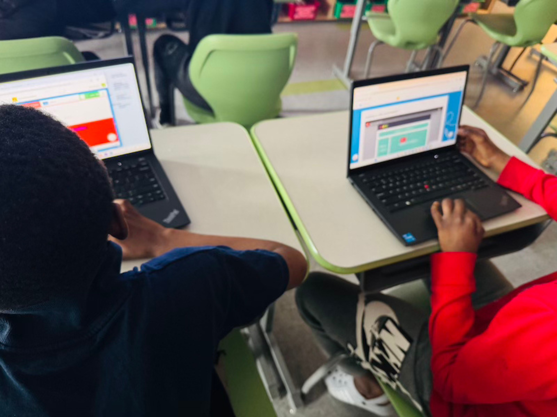 Two children working on laptops at a school desk in a classroom.