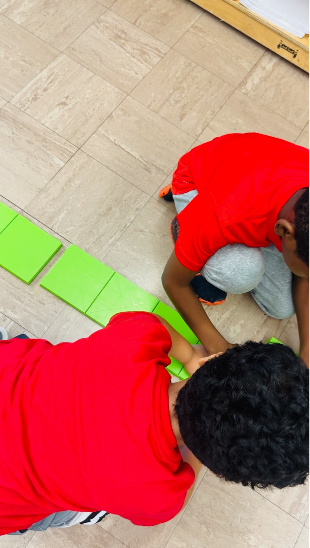 Two boys in red shirts building with green tiles on the floor.