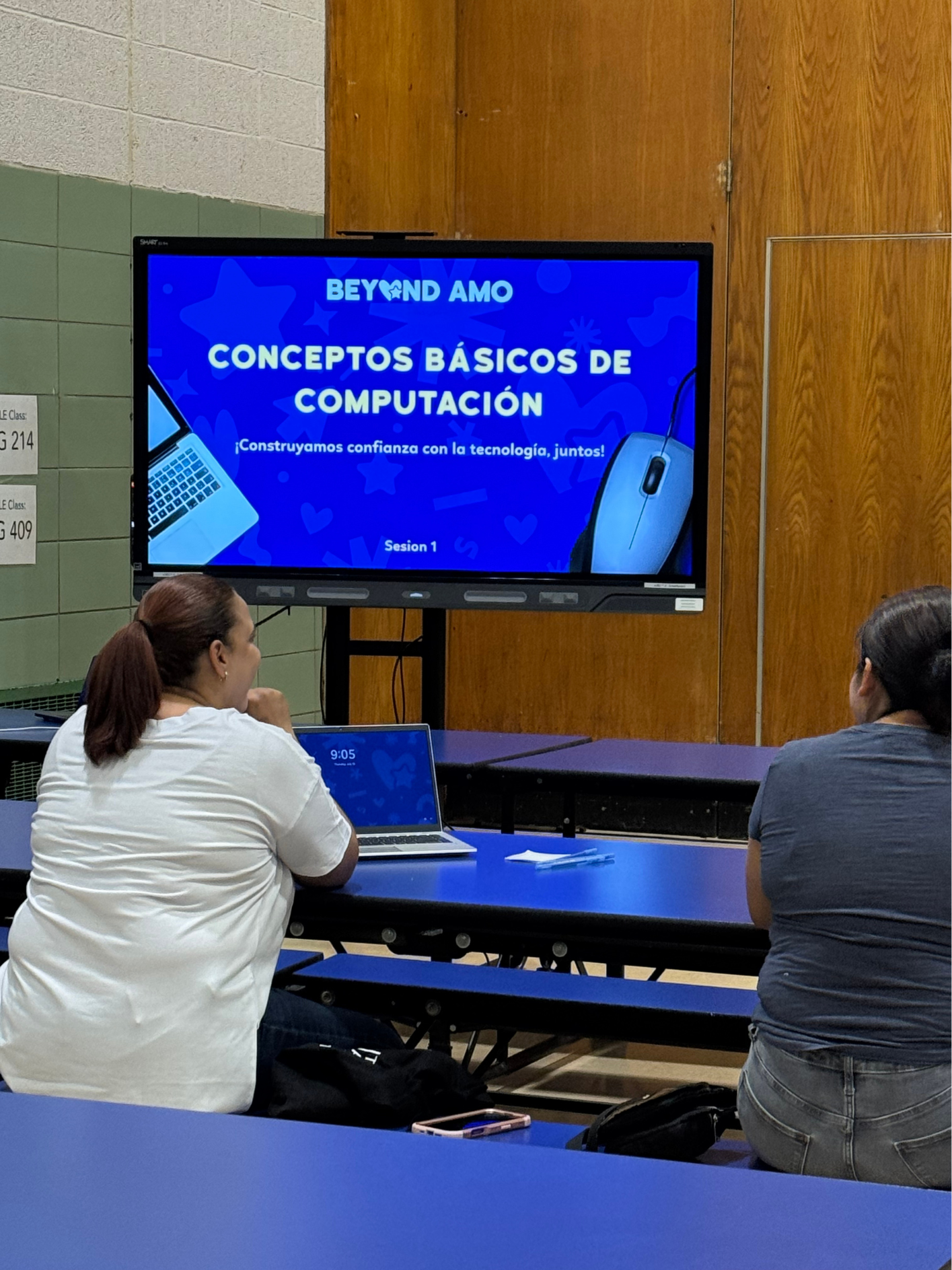 Two women sitting at a table in a classroom or conference room, facing a large screen displaying a presentation titled 'Conceptos Básicos de Computación' with a laptop and a computer mouse on the slide.