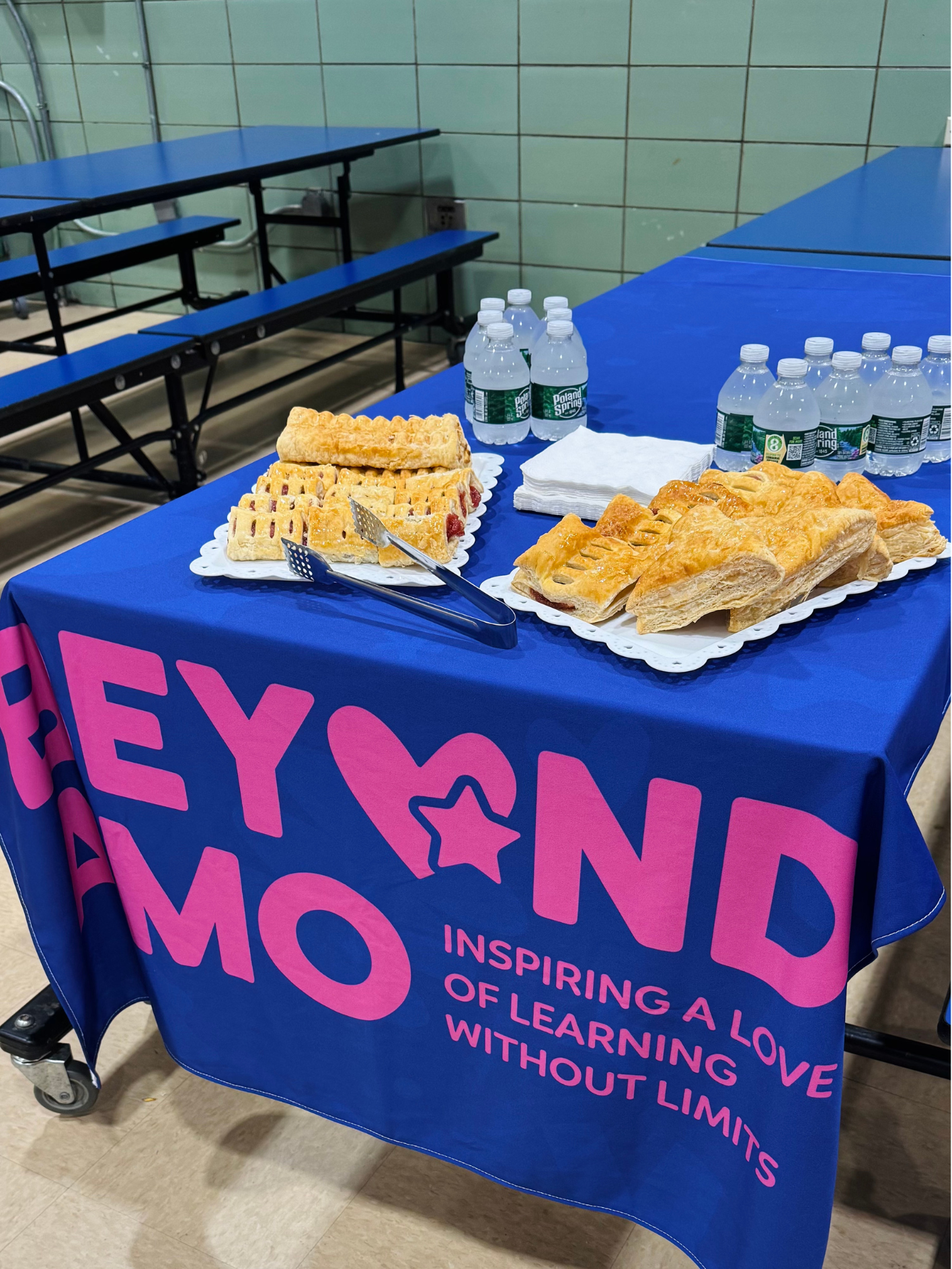 Table with water bottles, fruit pastries, and napkins at an event, with a purple tablecloth displaying the words "HERO AND MO" in pink letters and the phrase "Inspiring a love of learning without limits."