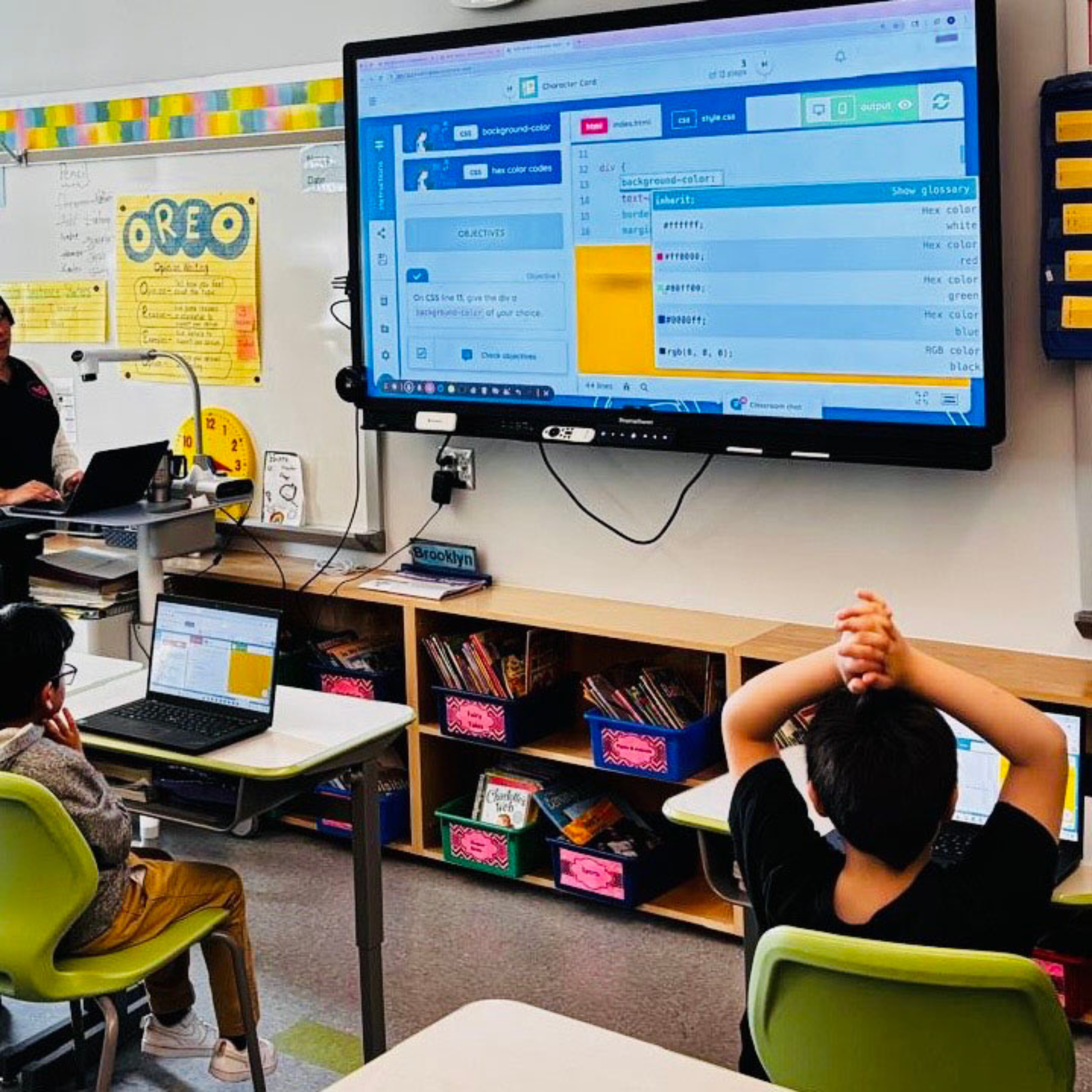 A classroom with students facing a large digital screen displaying a code editor, with one student in the foreground taking notes and others working on laptops.