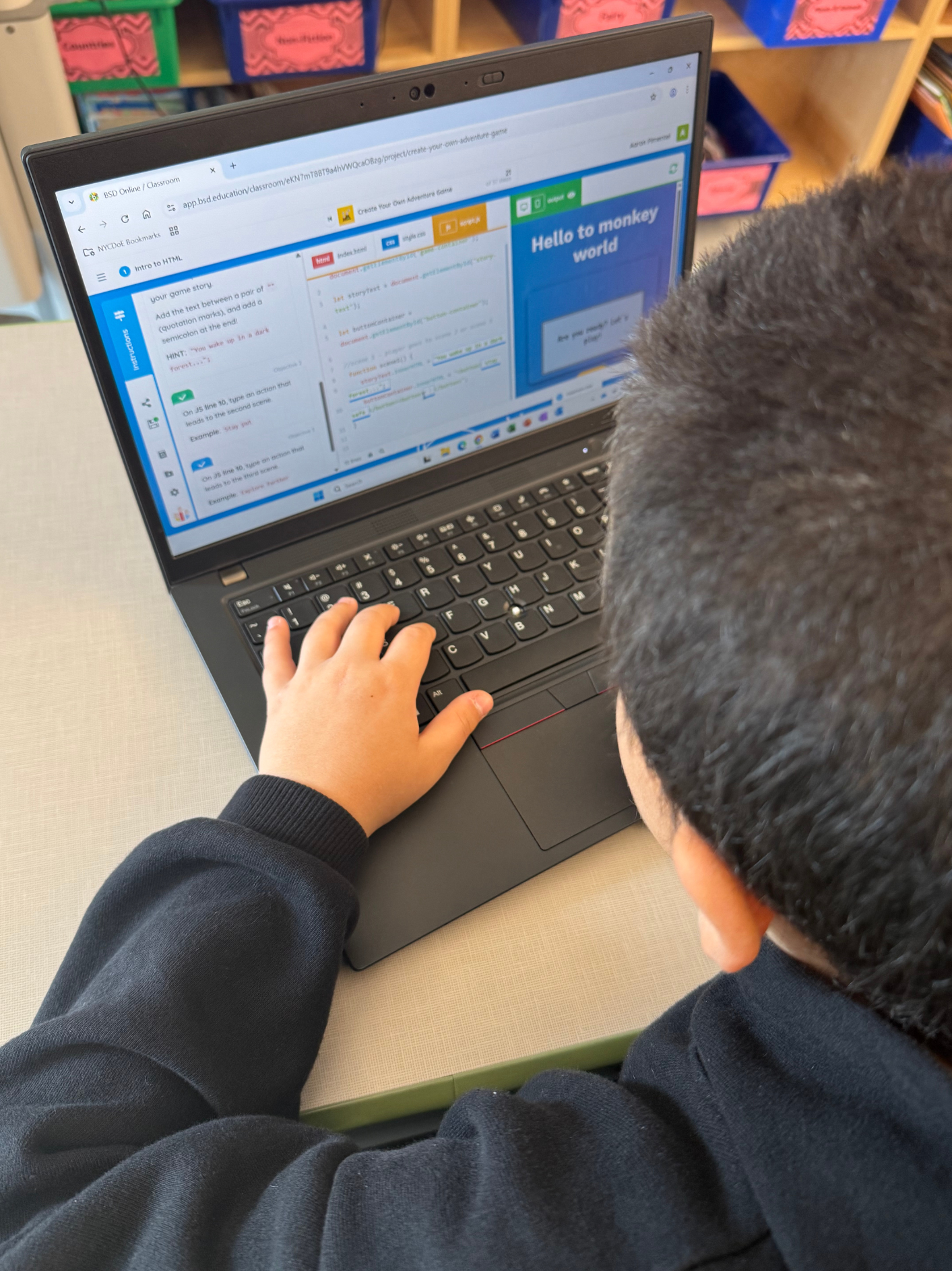 Child working on a laptop at a desk, displaying a coding or educational website with colorful sections and the text "Hello to monkey world".