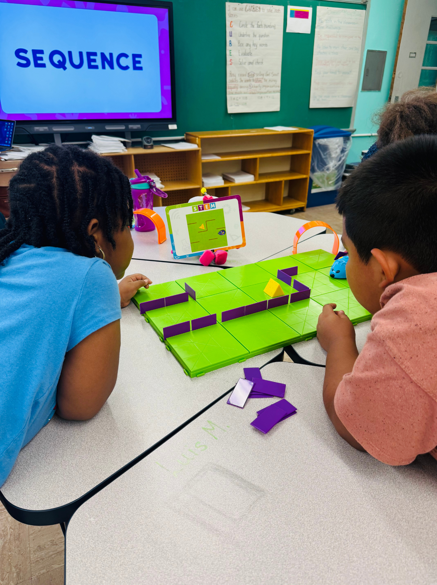 Two children playing a maze game with purple building pieces on a green game board, using a tablet displaying a maze app in a classroom.