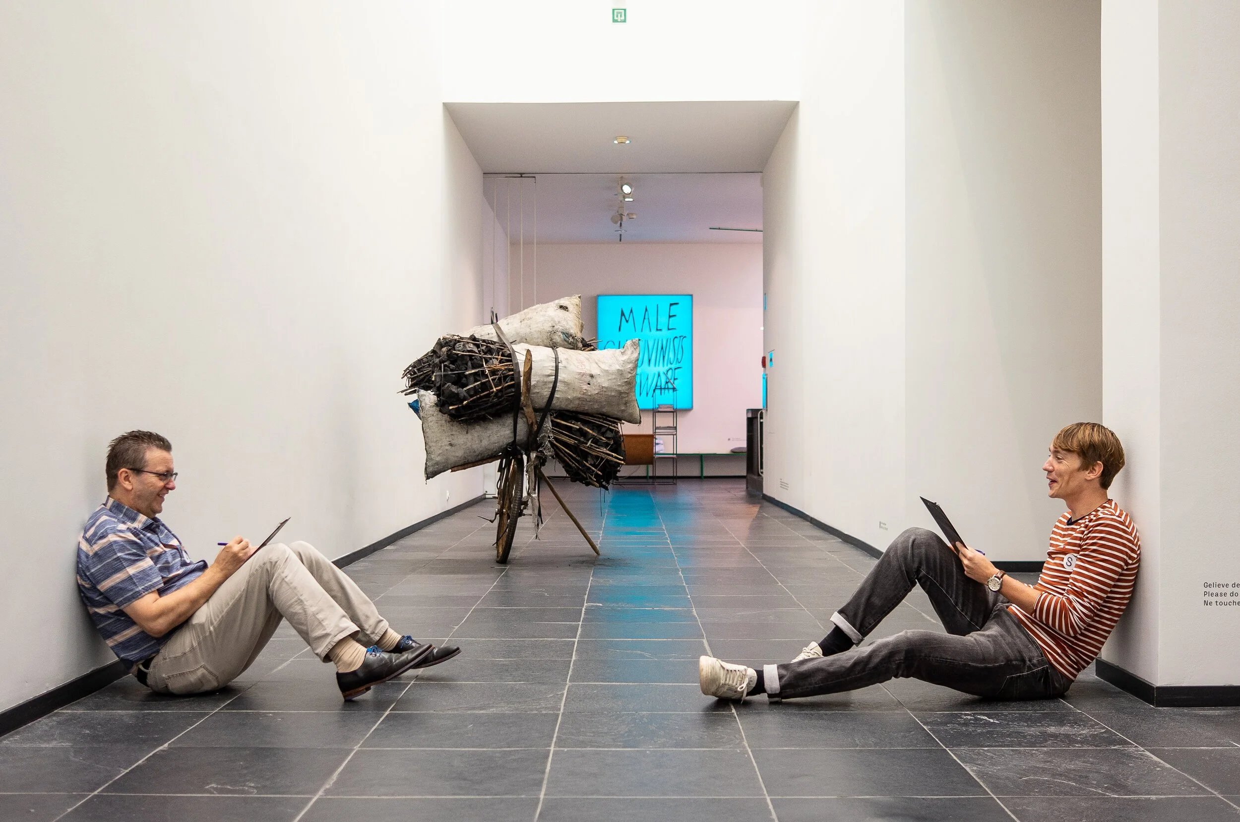 Two men sitting on the floor, leaning against white walls, smiling and looking at tablets, with an art installation on a bike between them. The installation has large fabric and wood-like objects. A digital screen in the background reads 'MALE', 'S',