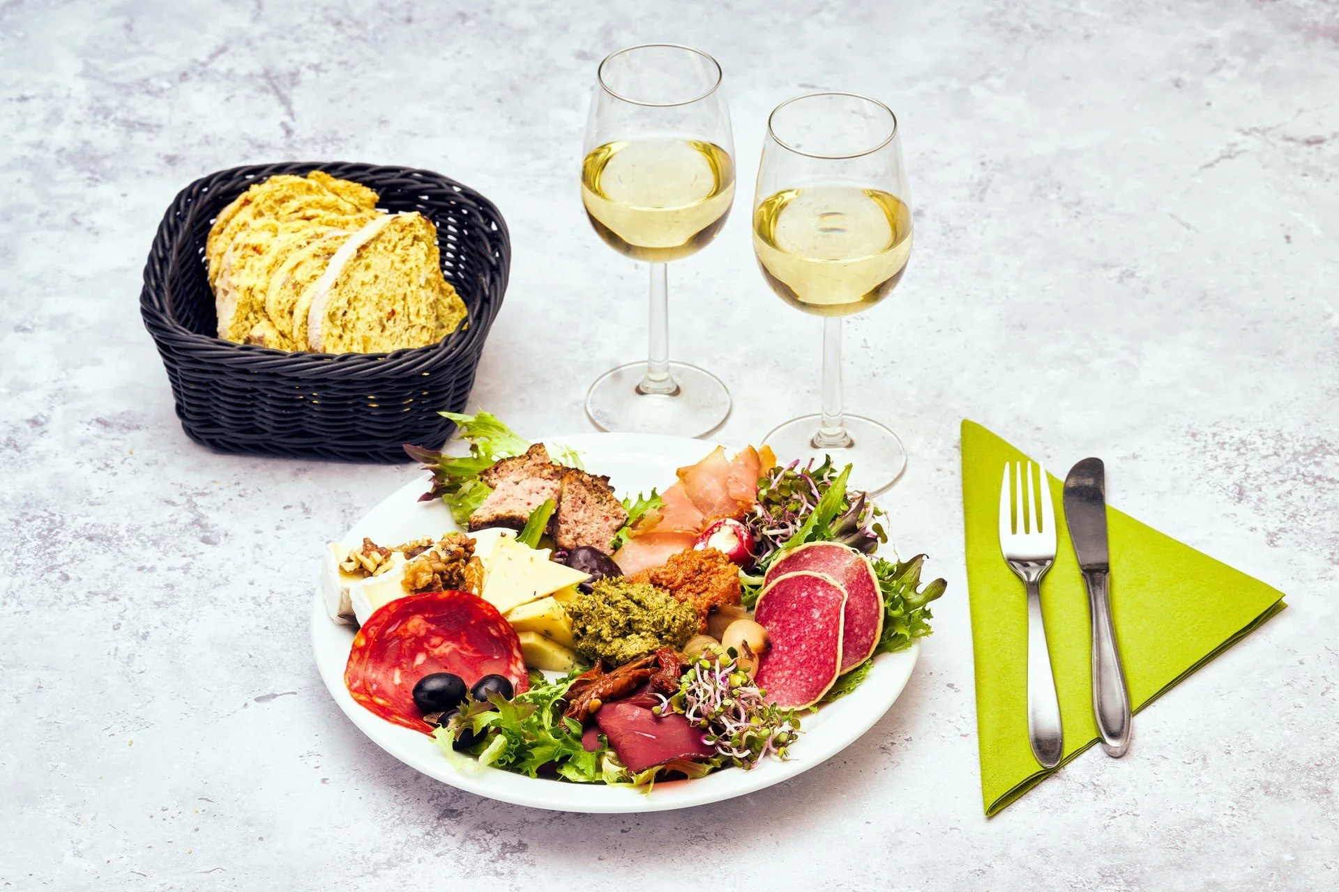 En tallerken med forskellig salat, skinke, ost og kød, to glas hvidvin, en skål med brødskiver, en grøn stofserviet med bestik.