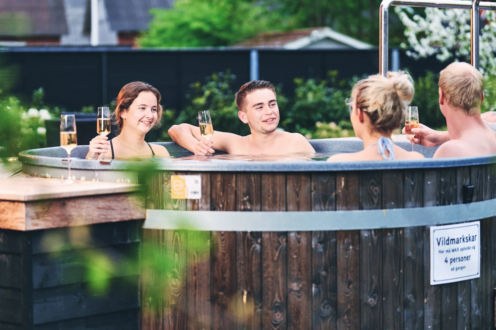 Fire personer nyder et afslappende bad i en udendørs hot tub, med tre af dem, der holder champagneglas, i en grøn have.