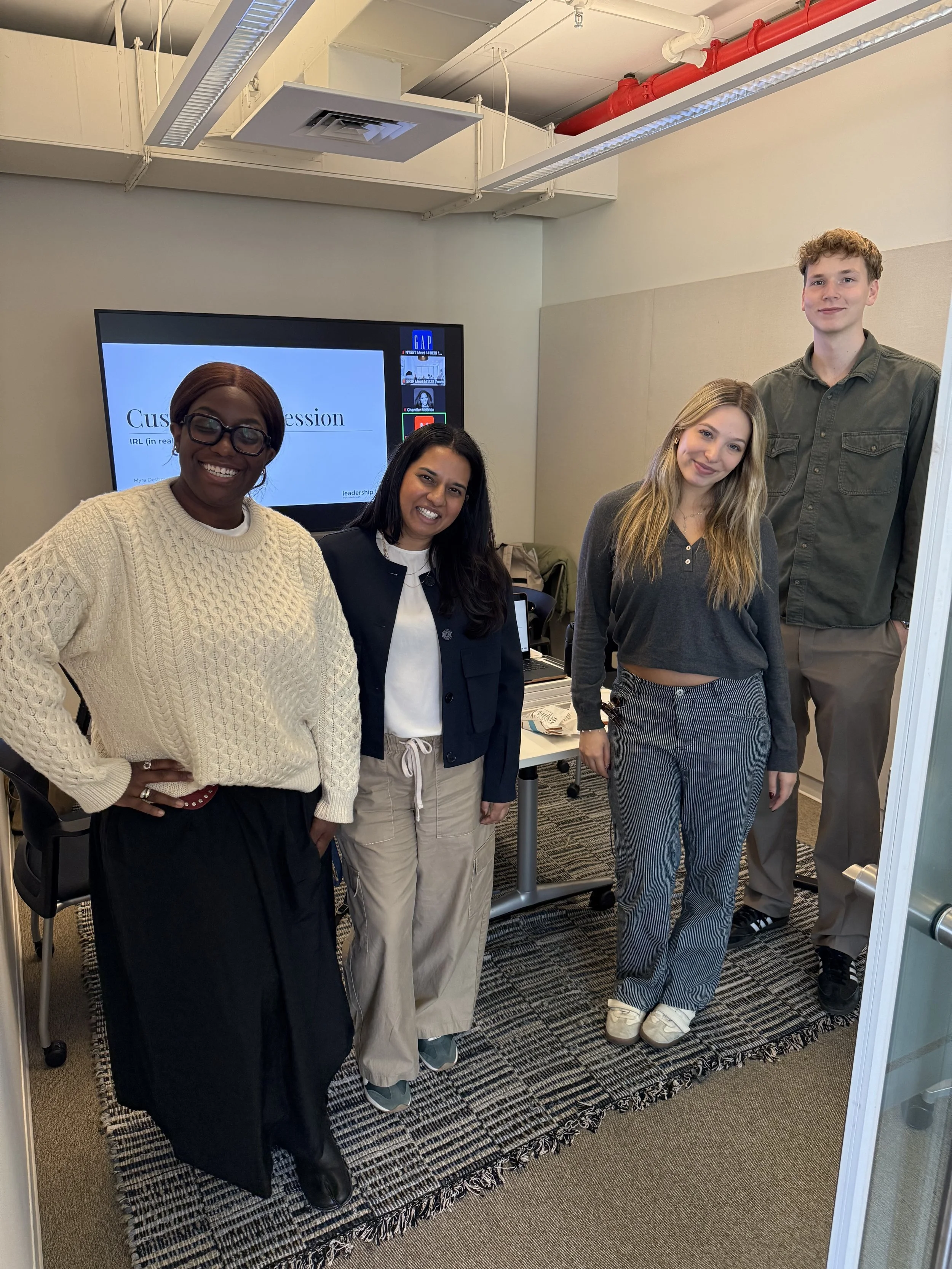 Four people standing in a conference room, smiling at the camera, with a presentation slide on a screen behind them.