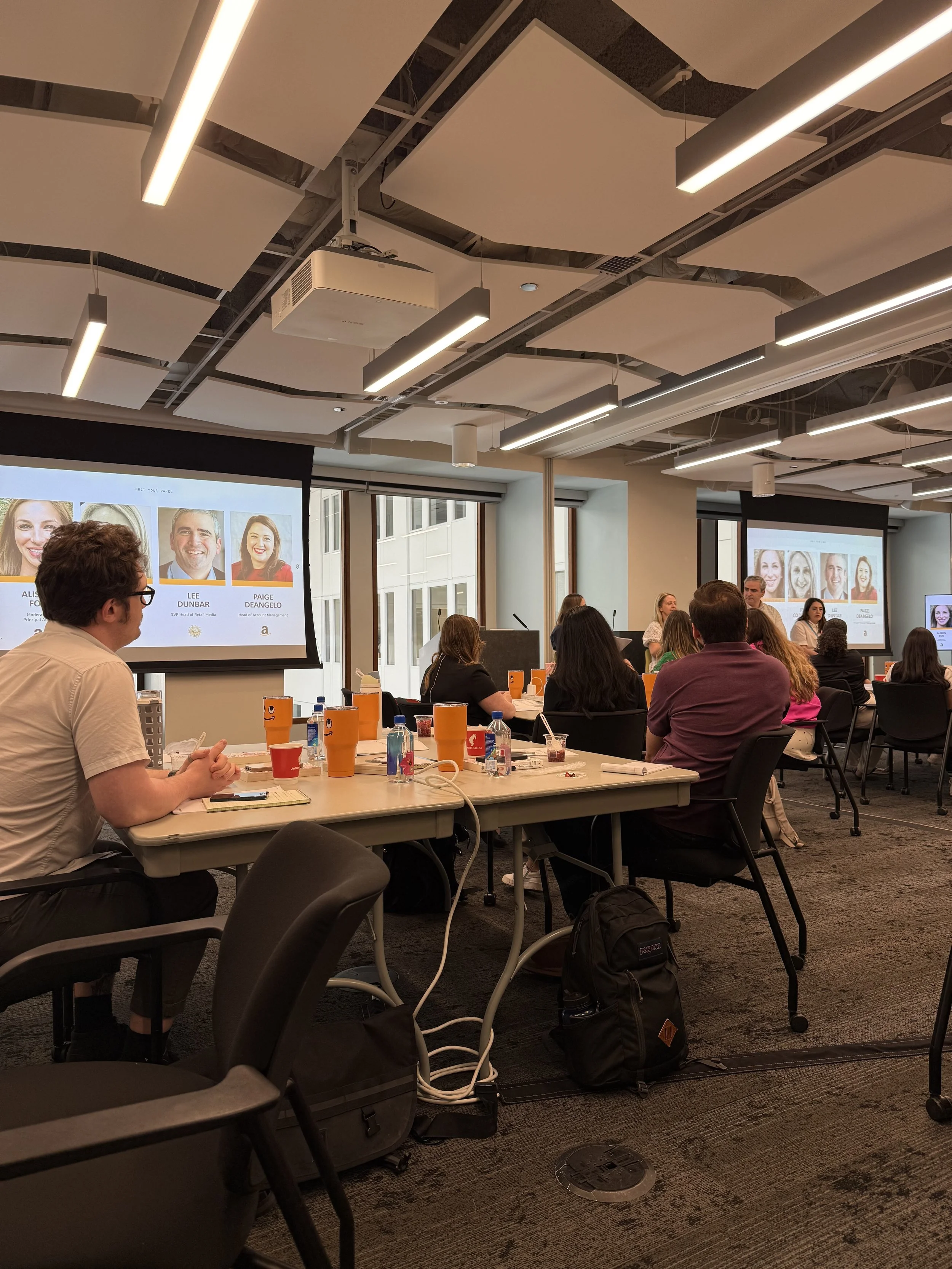 A conference room filled with people attending a presentation. There are two large display screens showing slides with headshots and names of speakers. The room has large windows and ceiling lights, with tables holding drinks, notebooks, and phones. 