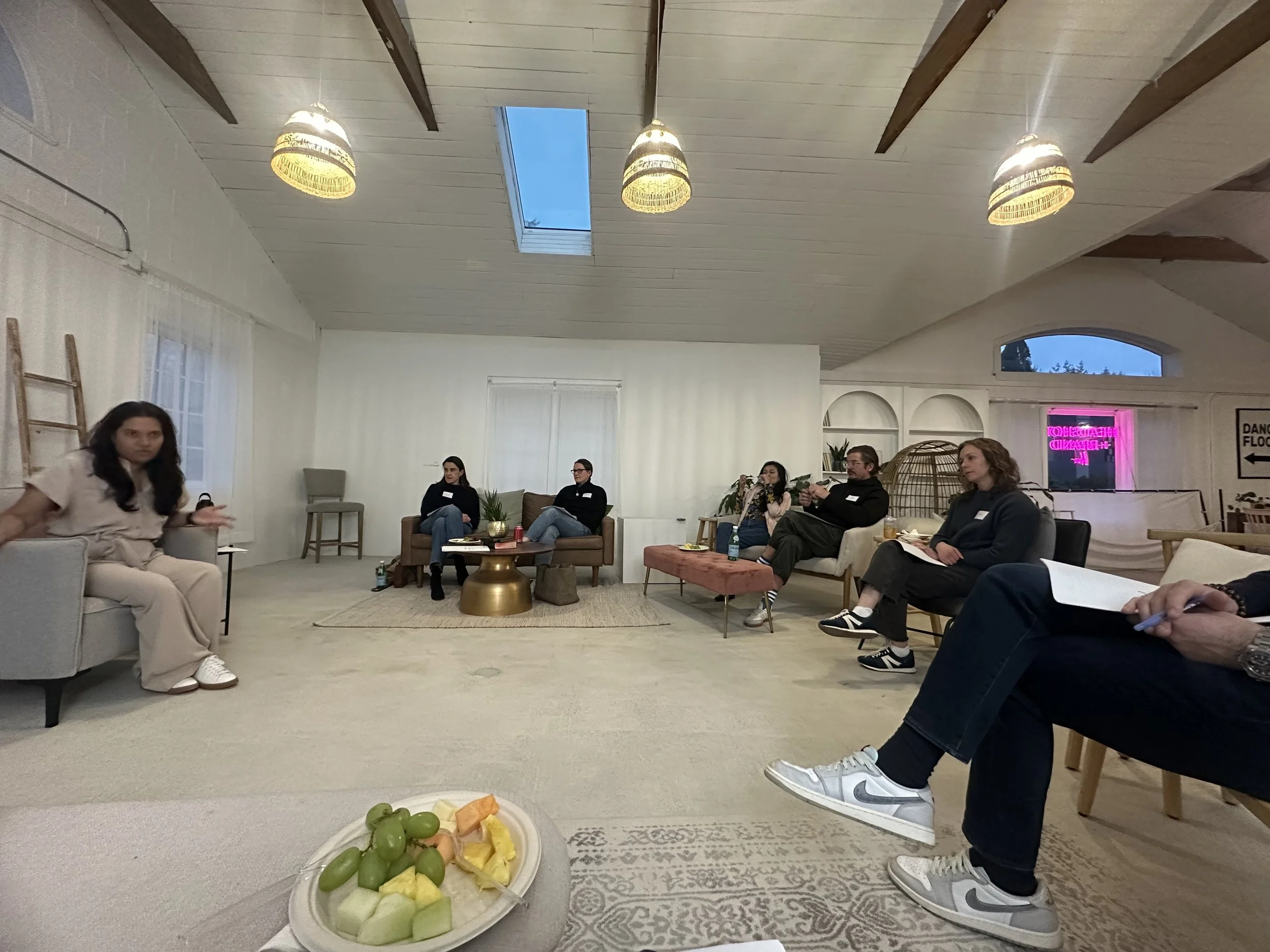 Group of people sitting in a spacious, well-lit room with a high ceiling during a discussion or presentation. There are seven individuals seated on various chairs and sofas, some taking notes. A plate of fruit in the foreground and some decorative it
