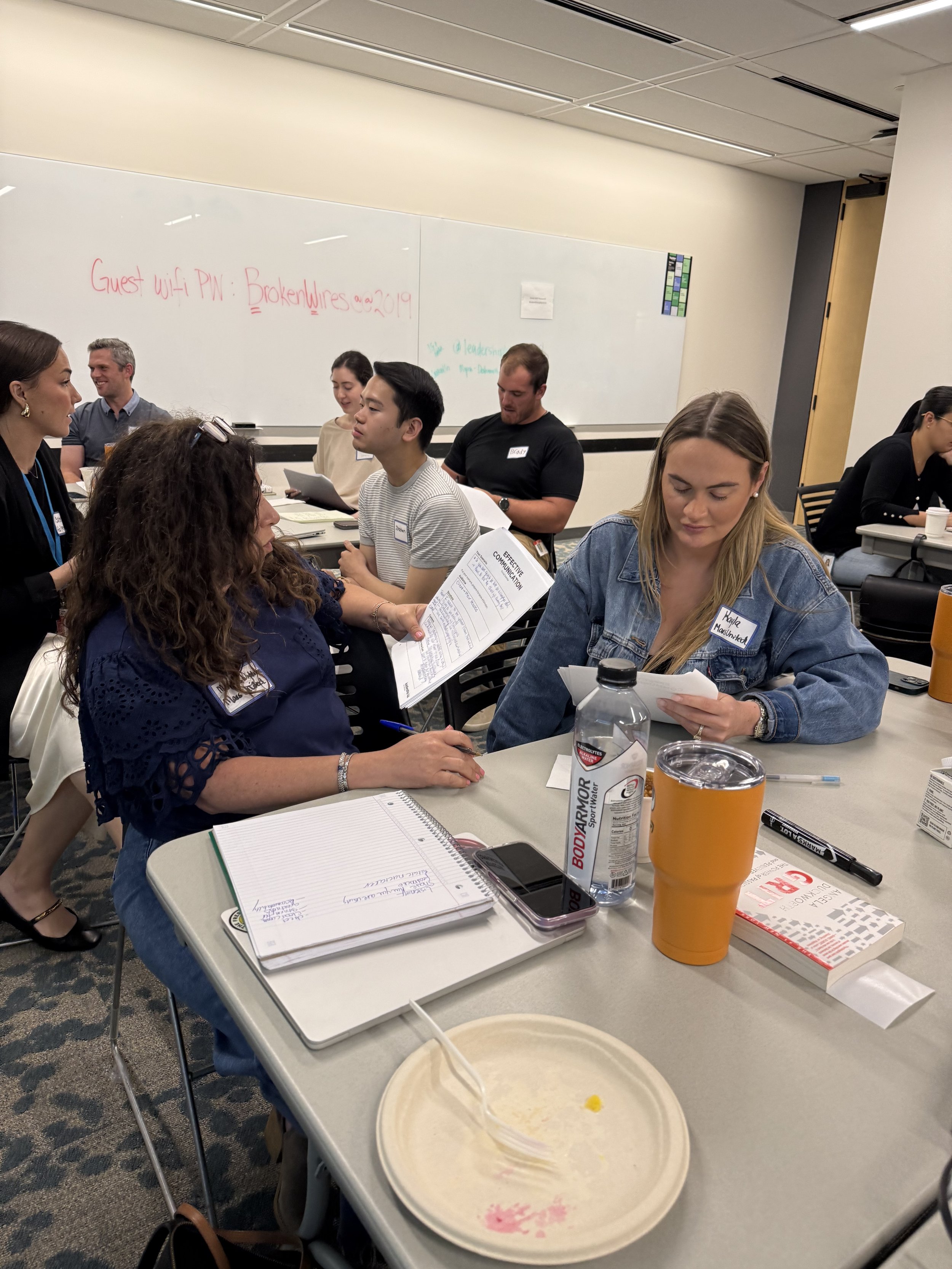 Group of people engaged in a discussion at a workshop in a classroom with whiteboards and see through plastic cups on the table.