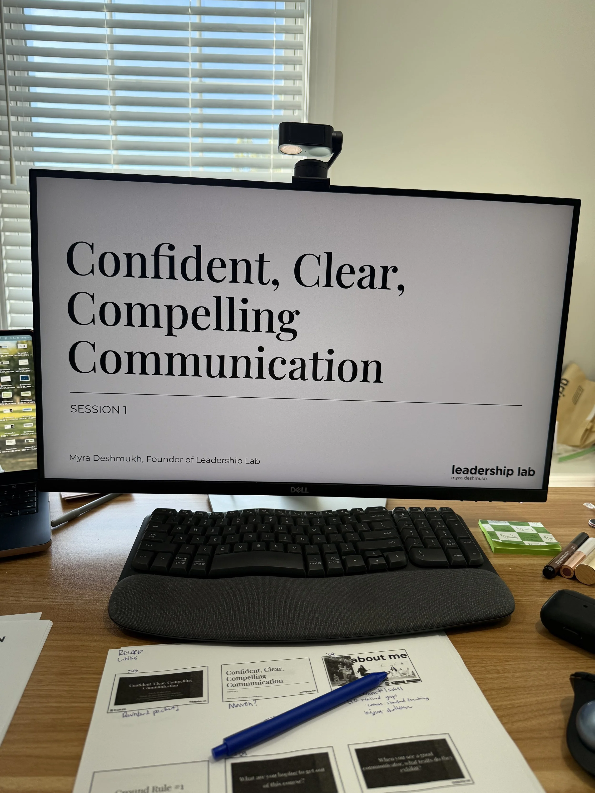 Computer monitor displaying a presentation titled 'Confident, Clear, Compelling Communication' by Myra Deshmukh on a desk with a keyboard, pen, papers, and office supplies.