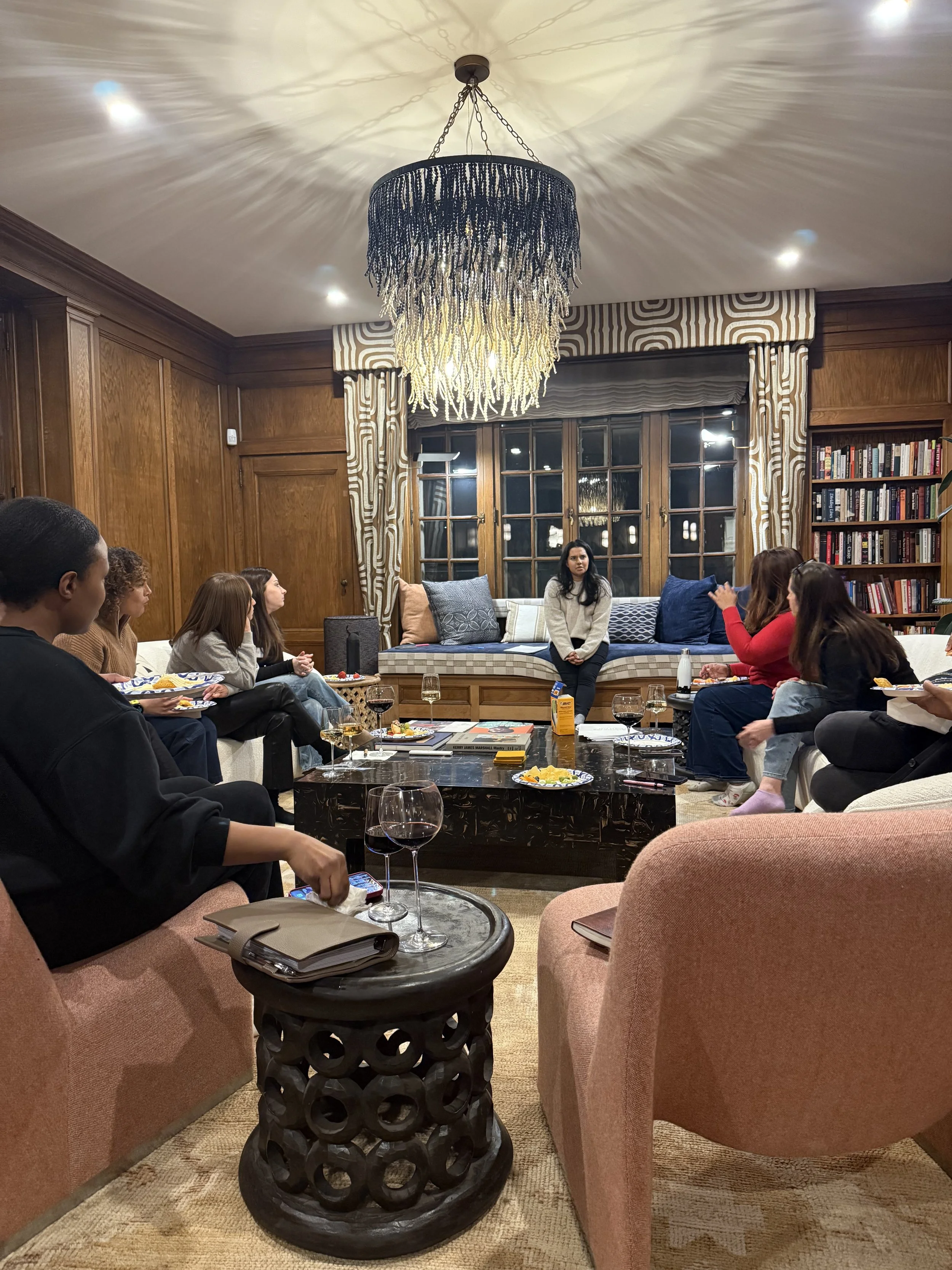 A group of people gathered in a living room, sitting on sofas and chairs, with one woman sitting on a built-in window seat. They are having a casual gathering with snacks and drinks, and one woman appears to be speaking or leading the discussion. The