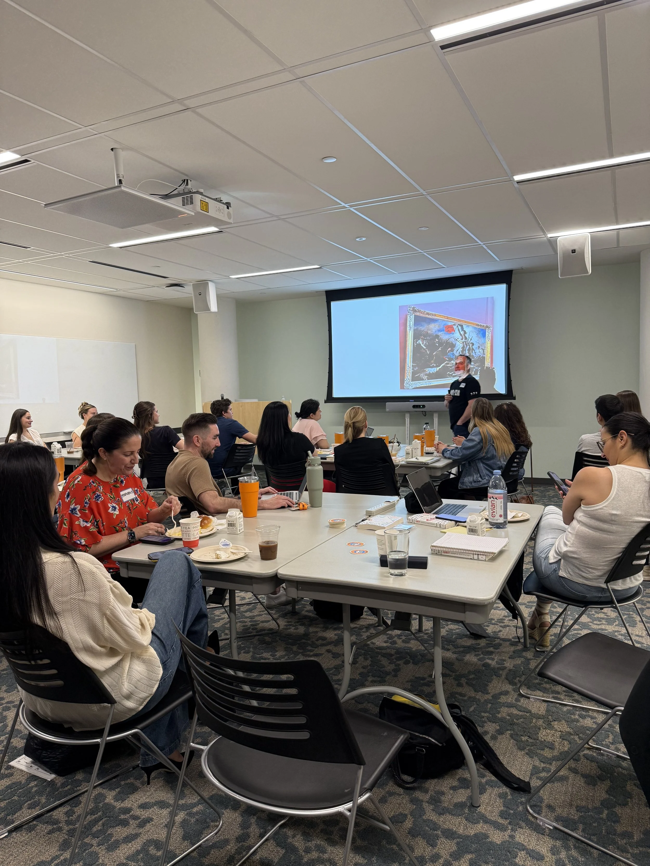 A classroom or conference room filled with adult students, some eating, some using laptops, listening to a speaker at the front, with a large screen displaying a colorful artwork.