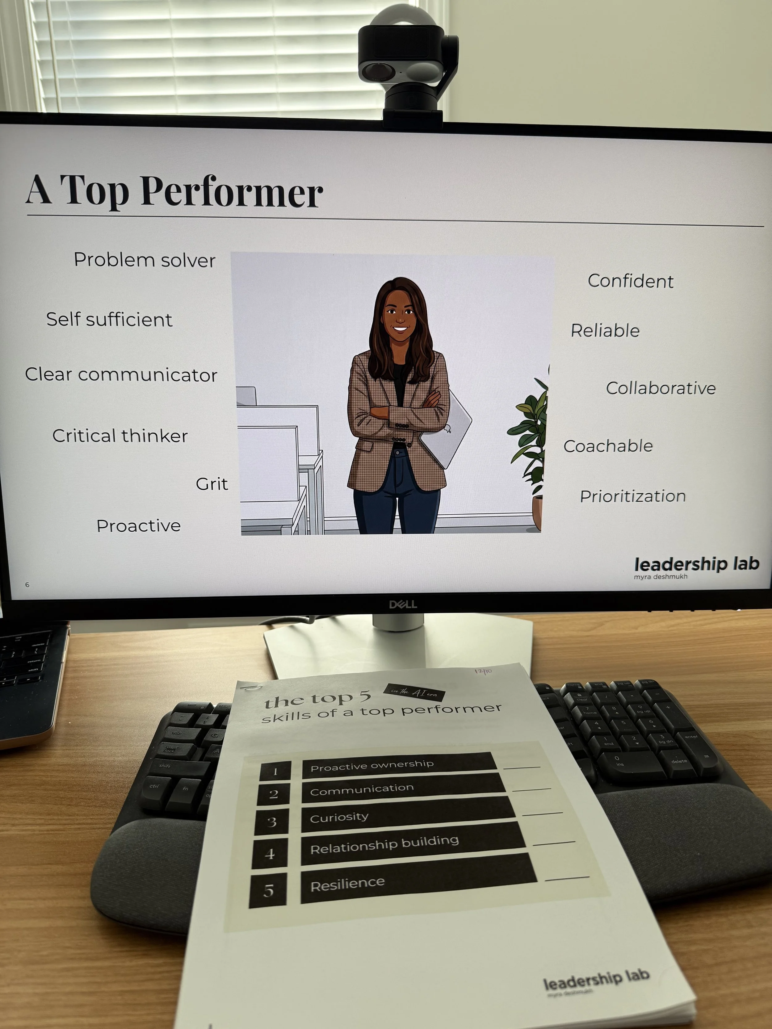 Computer monitor displaying a presentation slide titled "A Top Performer" with a cartoon woman with brown hair and a blazer, surrounded by words like "Problem solver," "Confident," and "Prioritization." A keyboard and a printed top 5 skills list are 