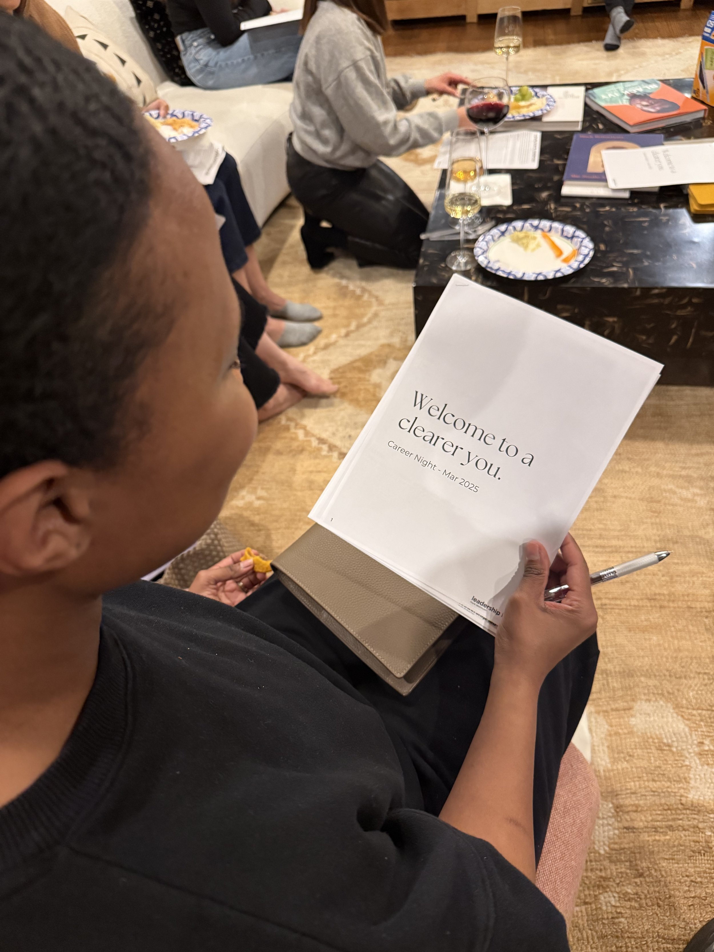 Person holding a booklet titled 'Welcome to a clearer you. Career Night - Mar 2025' during a group event, with a table of food and drinks visible in the background.