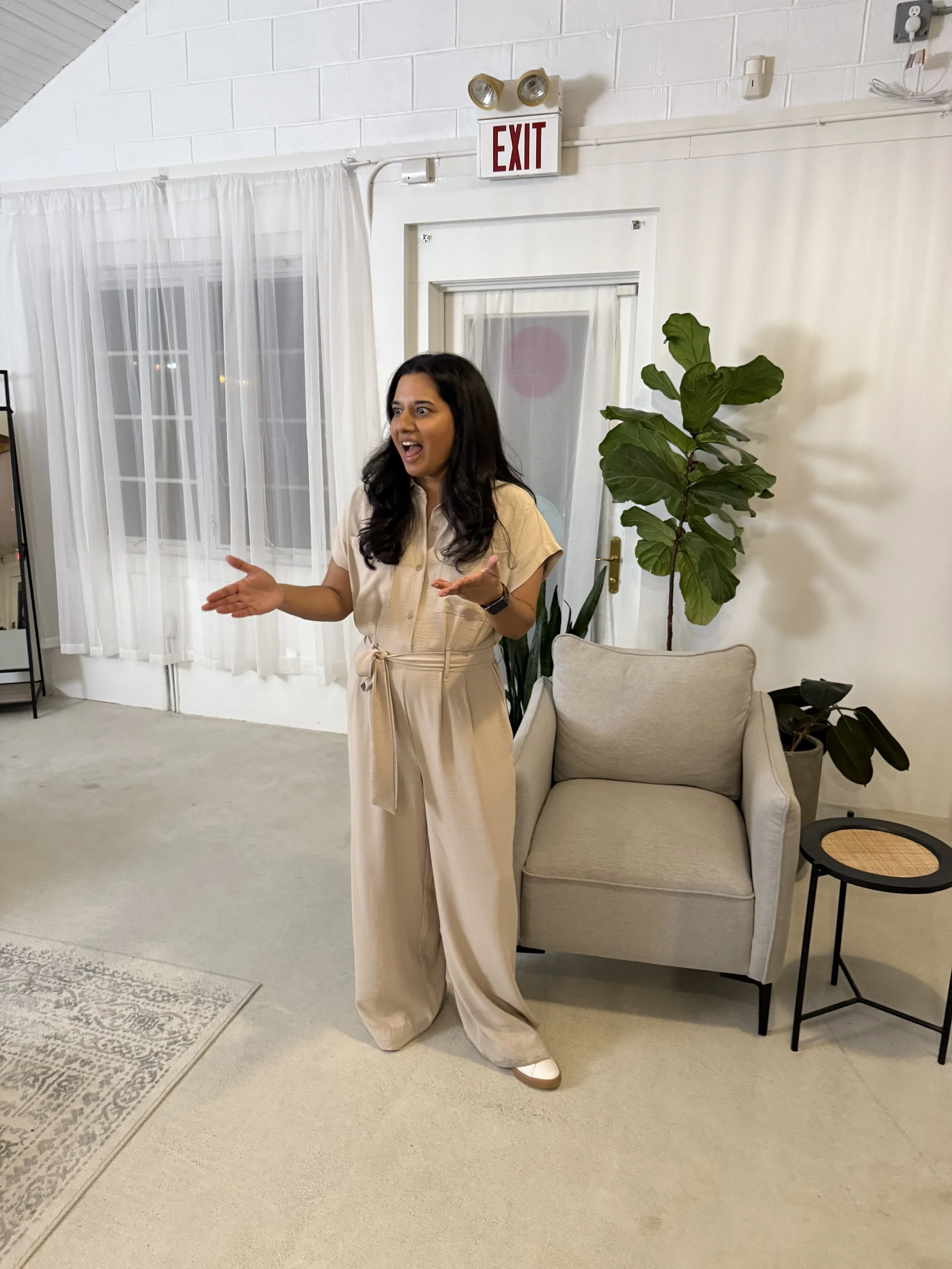 Woman standing indoors, gesturing with hands, talking or explaining, near a beige armchair and a tall plant, in a room with white brick walls and white curtains.