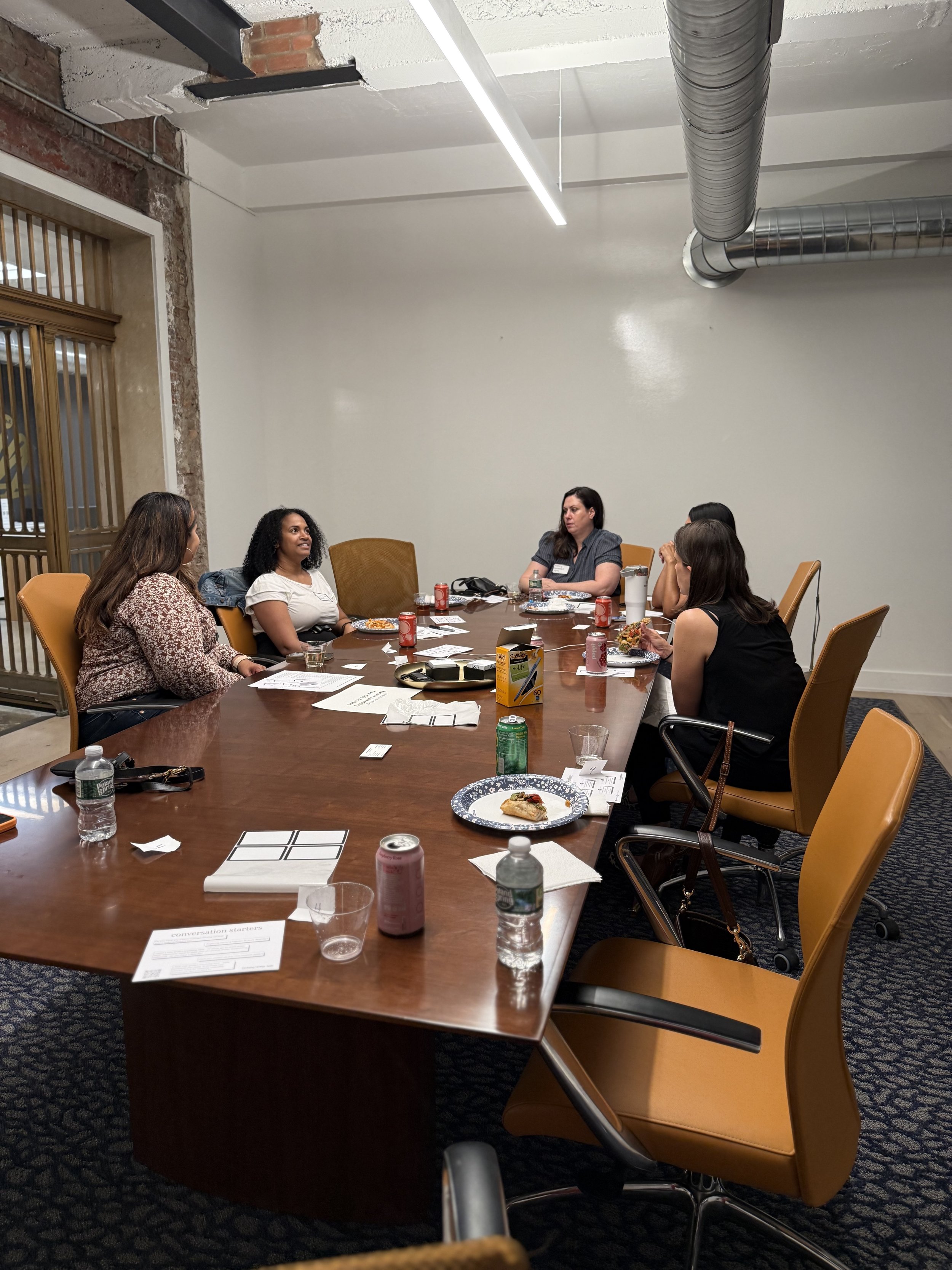 A group of five women seated around a conference table in a meeting room, engaged in conversation.