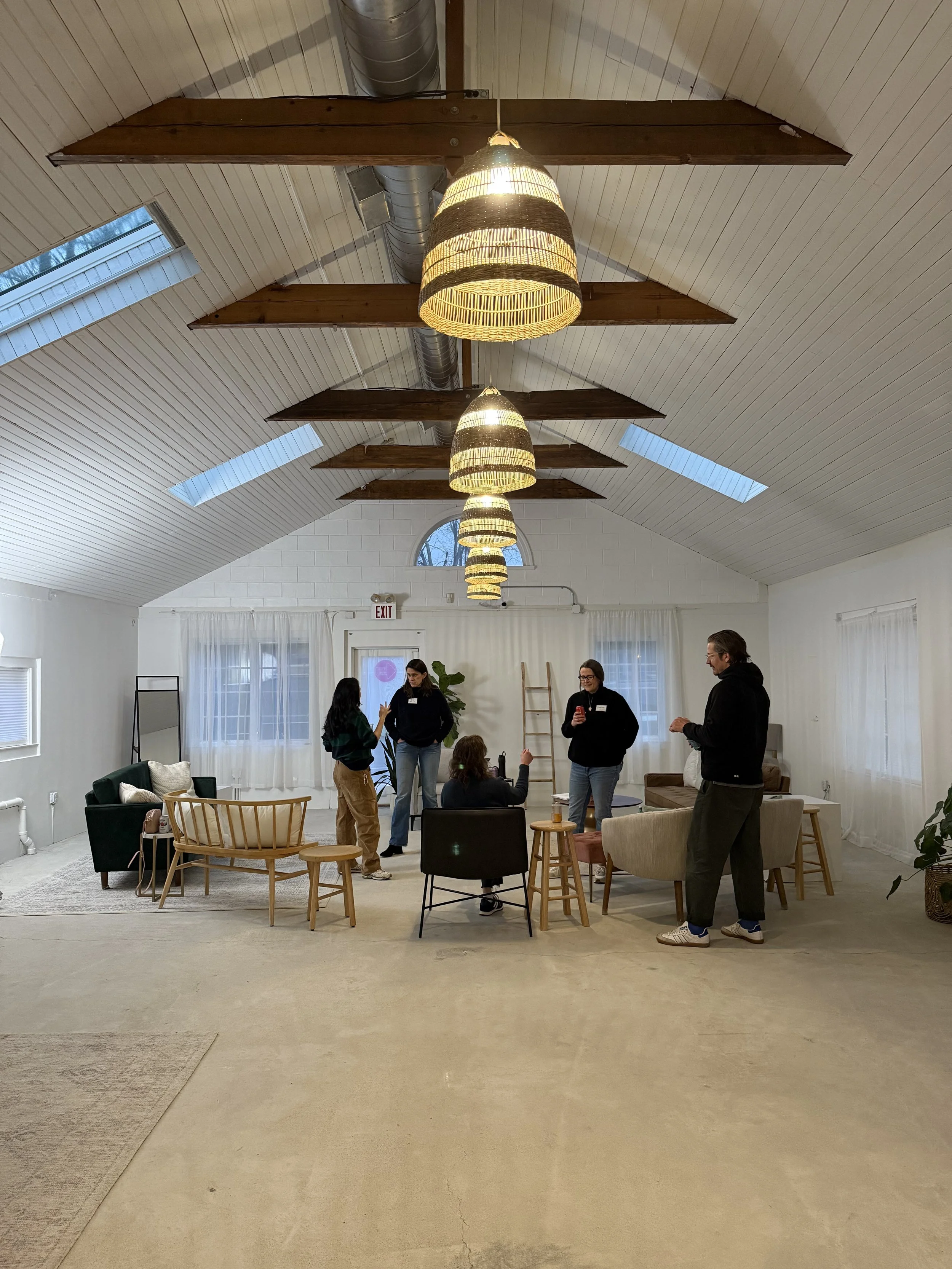 Group of people talking and sitting in a well-lit, modern room with high vaulted ceilings, hanging lamps, and natural light from skylights and windows.