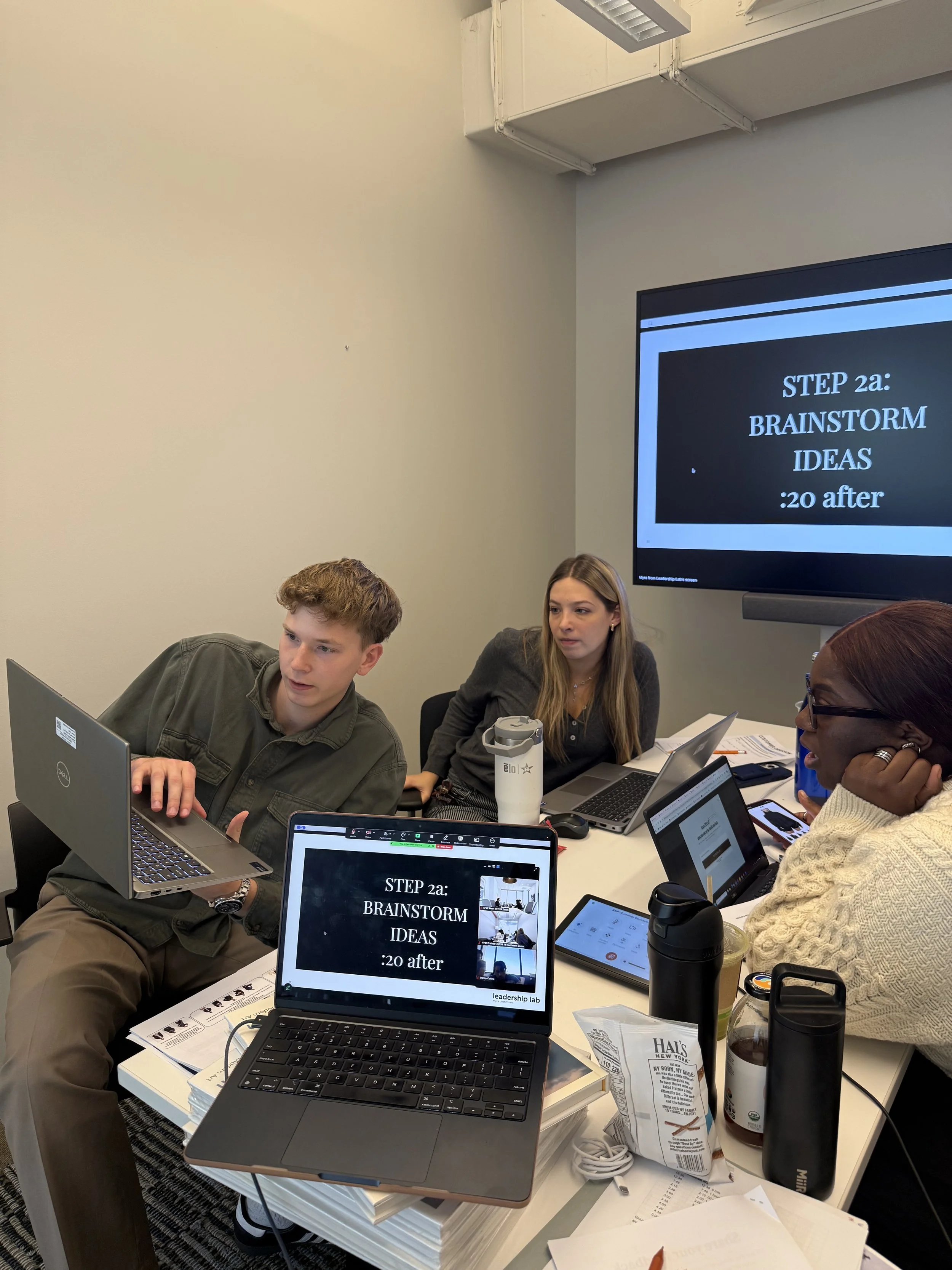 Group of three people in a meeting room with laptops, tablets, and notebooks, participating in a brainstorming session. Two laptops display a slide with the text "STEP 2a: BRAINSTORM IDEAS :20 after". There are papers, drinks, and personal items on t
