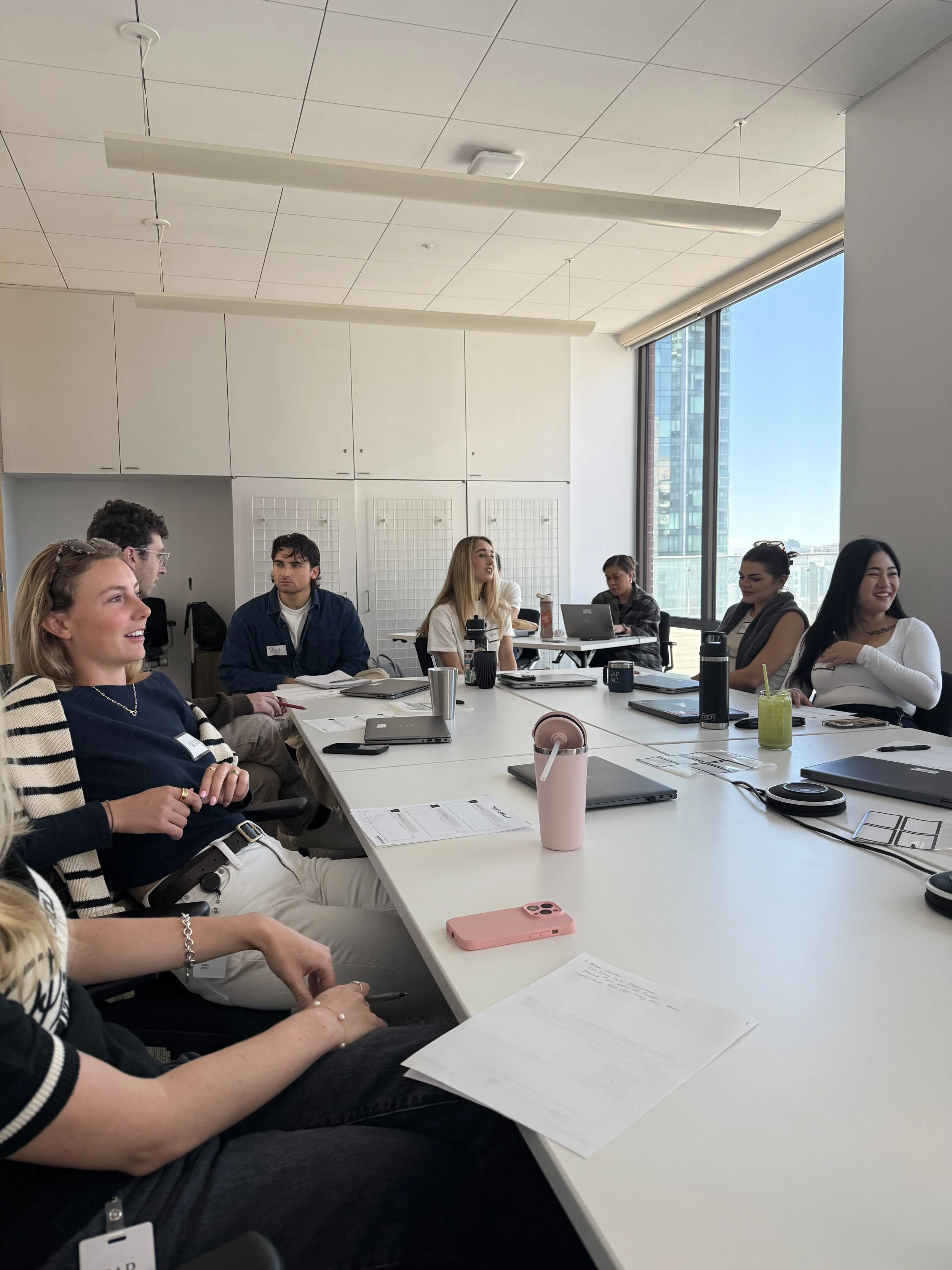 Business meeting with multiple people seated around a long conference table in a modern office, with laptops, notebooks, and drinks, and a large window showing city buildings outside.