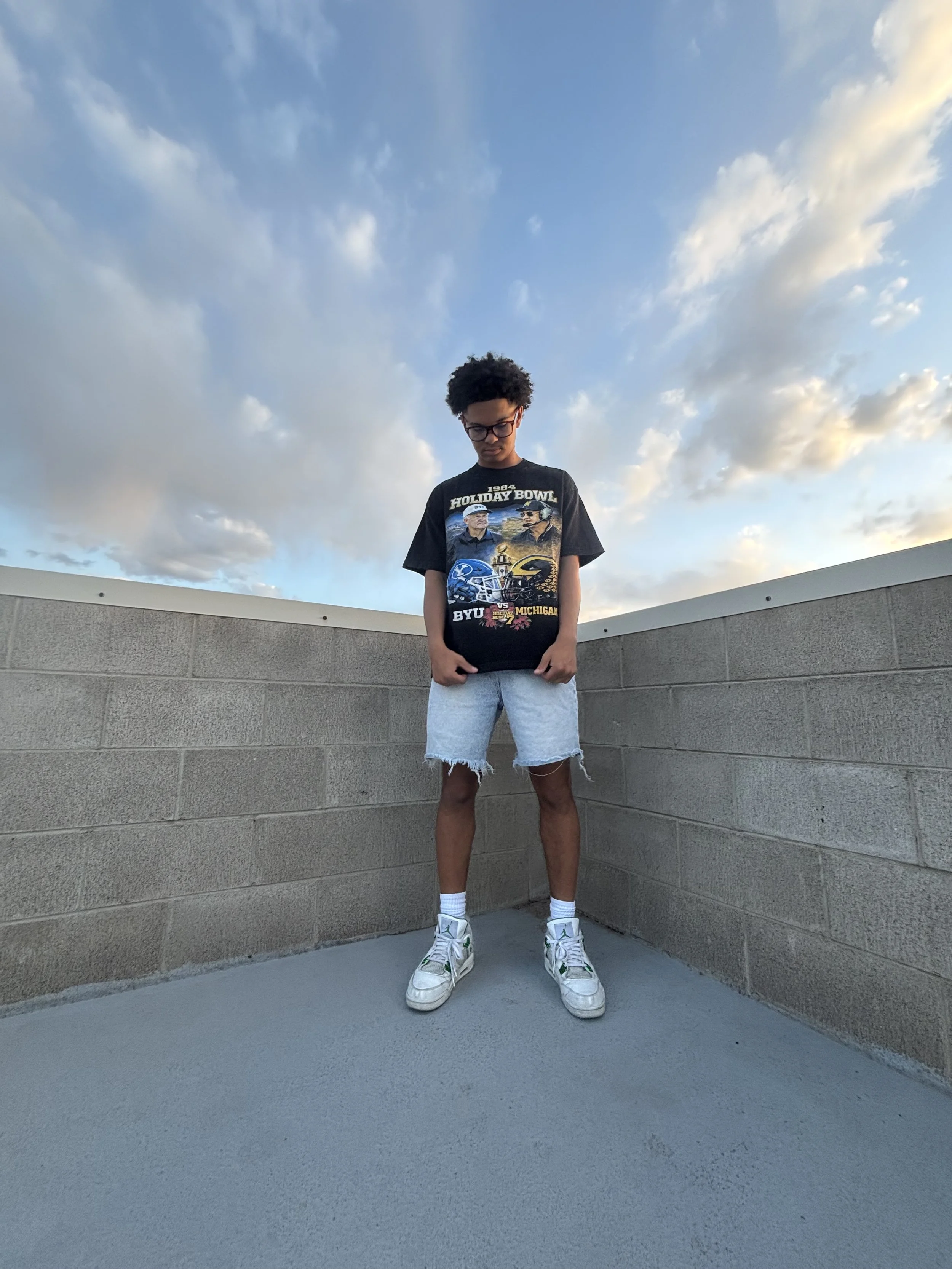 A young man with curly hair and glasses standing against a gray brick wall with a partly cloudy sky above, wearing a black T-shirt with football team graphics, white shorts with frayed edges, white socks, and sneakers.
