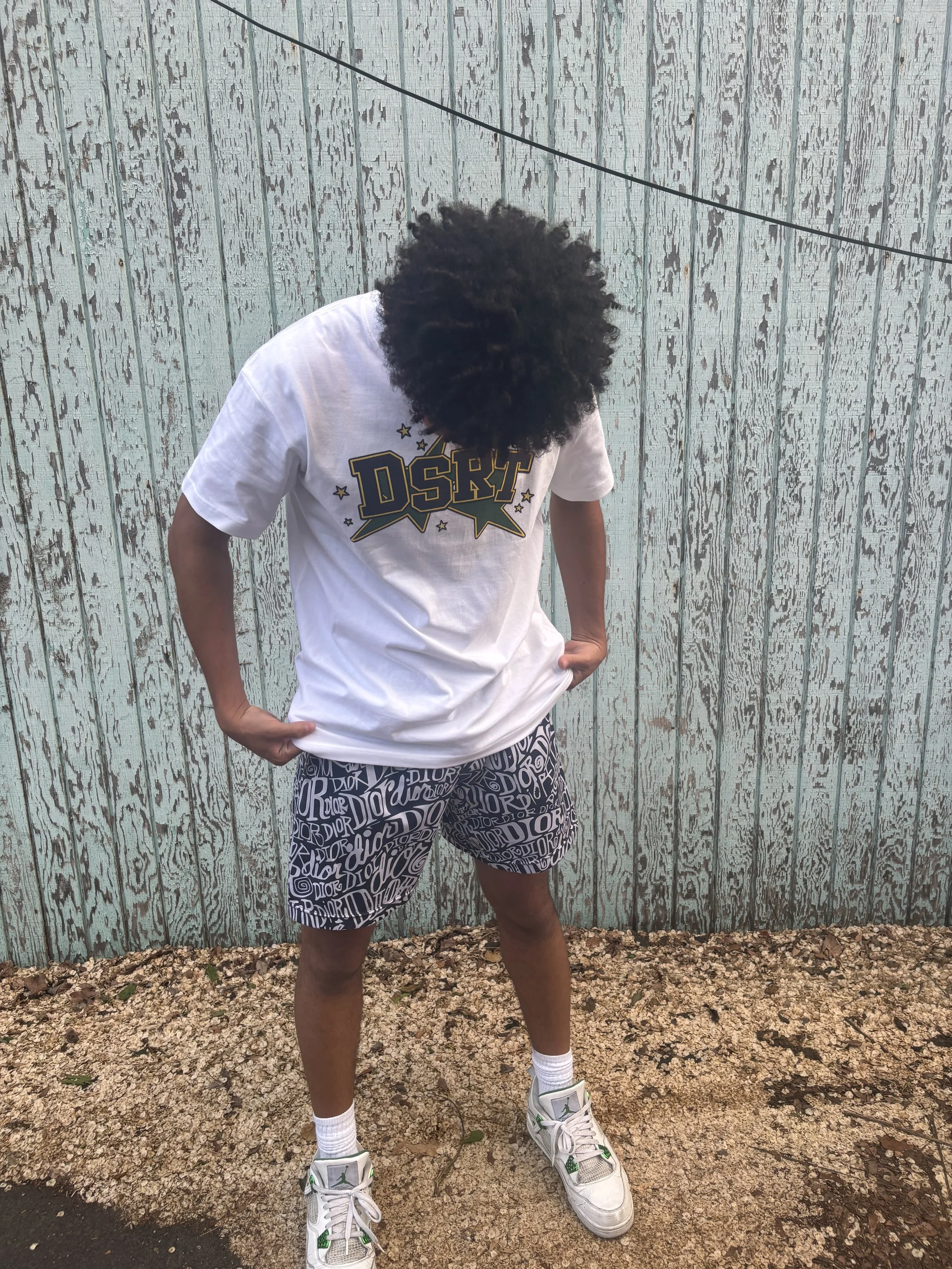 Person standing outdoors in front of a weathered wooden fence, wearing a white t-shirt, patterned shorts, white socks, and sneakers with a Jumpman logo, looking down with hands on hips.