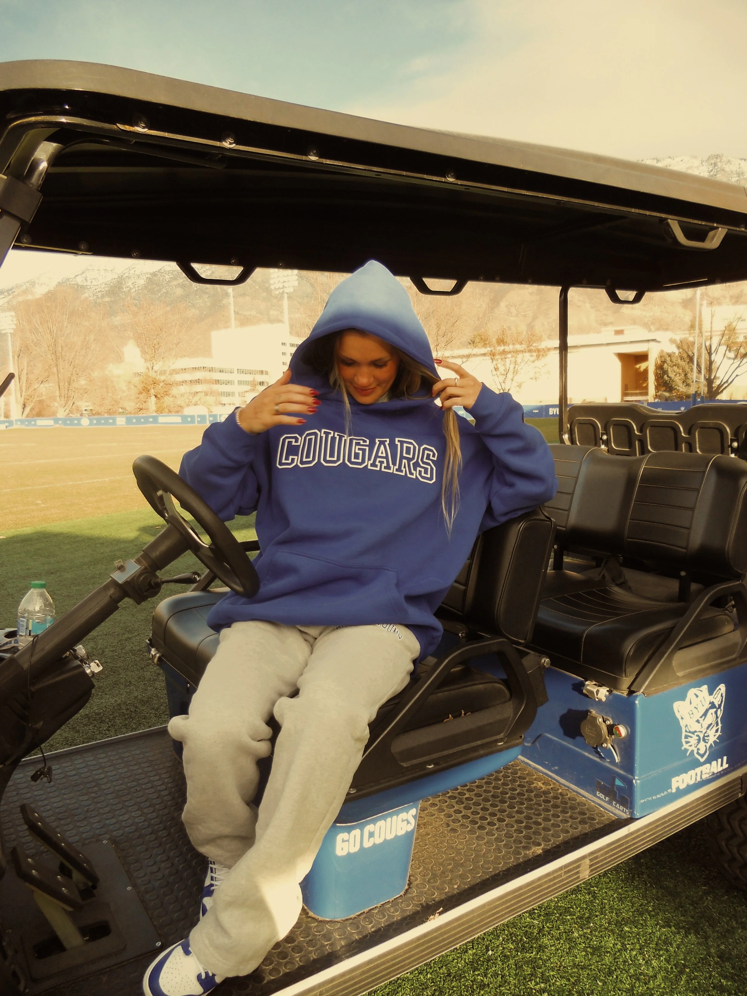 A woman sitting in a golf cart on a grassy field, wearing a blue hoodie with 'COUGARS' written on it and beige sweatpants, adjusting her hoodie.