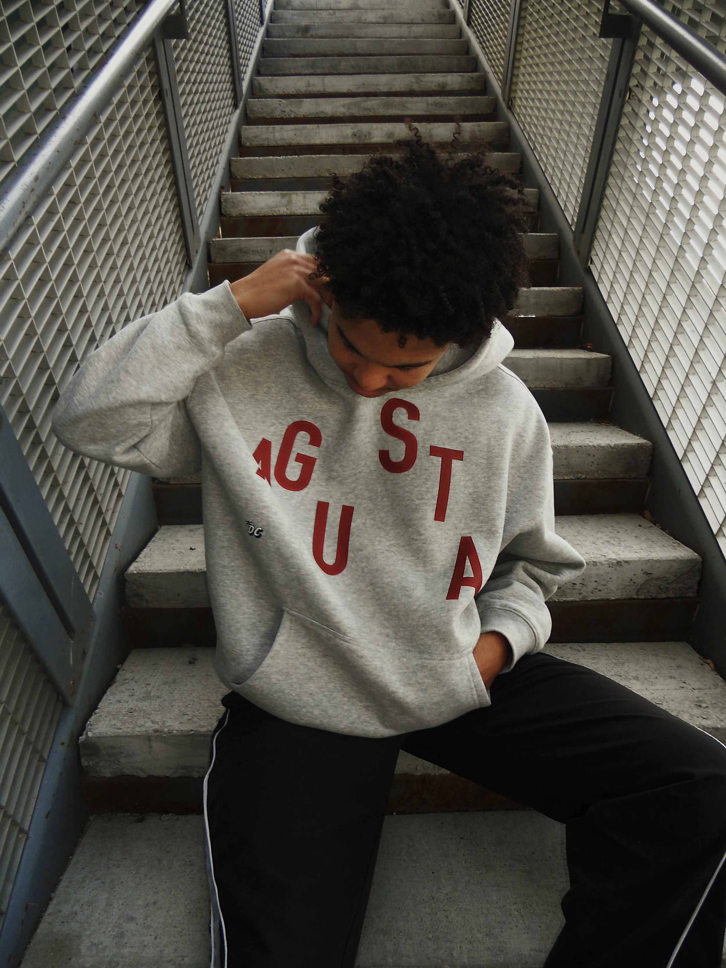 A young man with curly black hair, wearing a gray hoodie with red letters, sitting on outdoor stairs with a metal railing and lattice fencing on the sides, looking down and scratching his head.