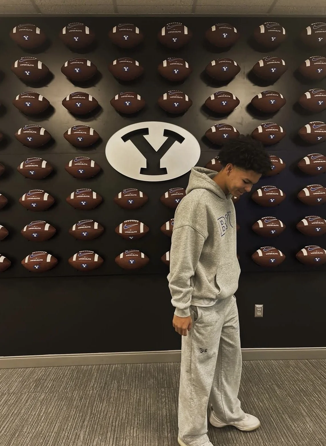 A young man in gray hoodie and sweatpants standing in front of a wall with many footballs and a large BYU logo.